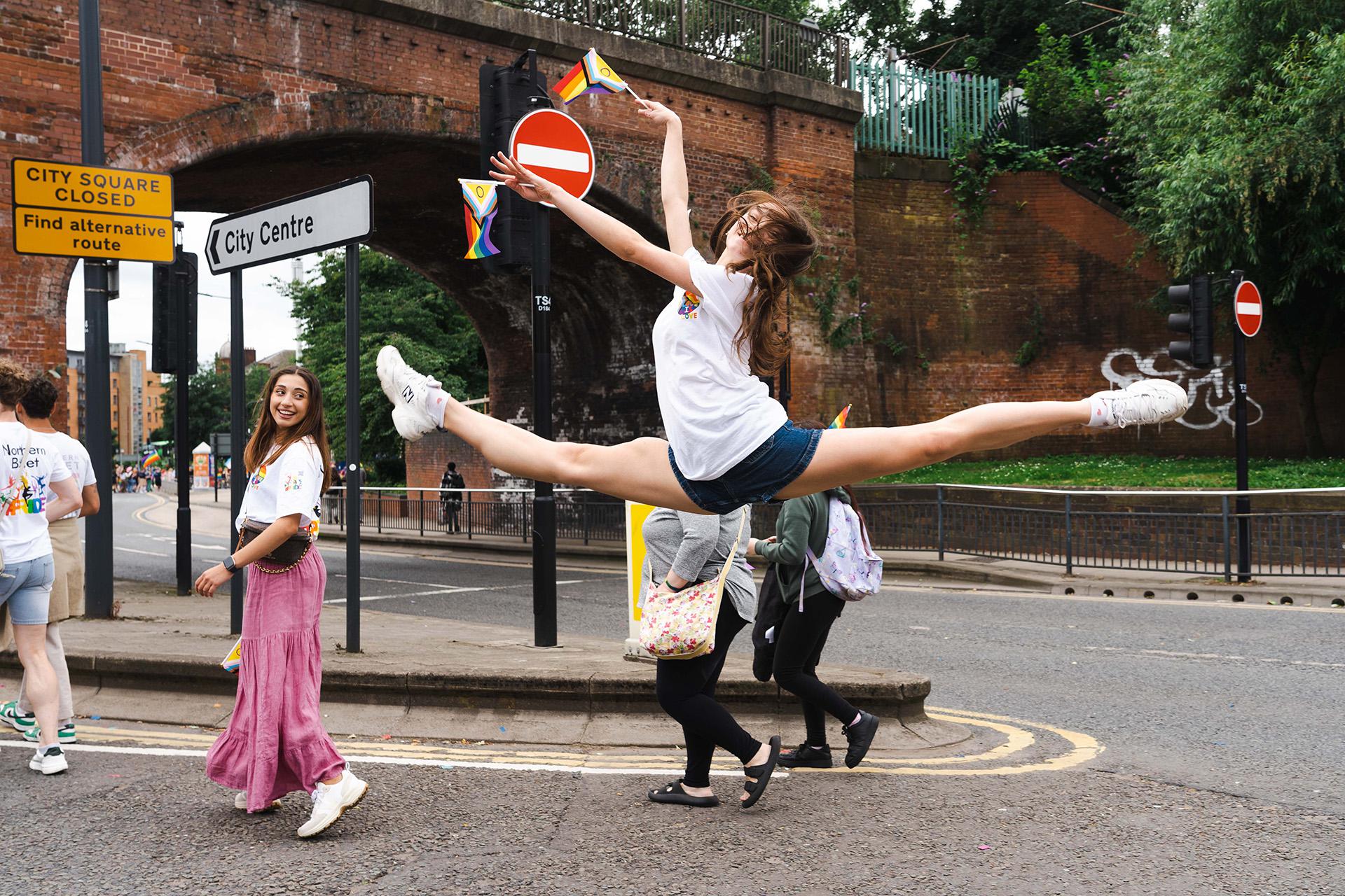 Dancer performing a grande jete wearing white strainer, denim shorts, a white T-shirt, and waving a Pride Inclusivity flag