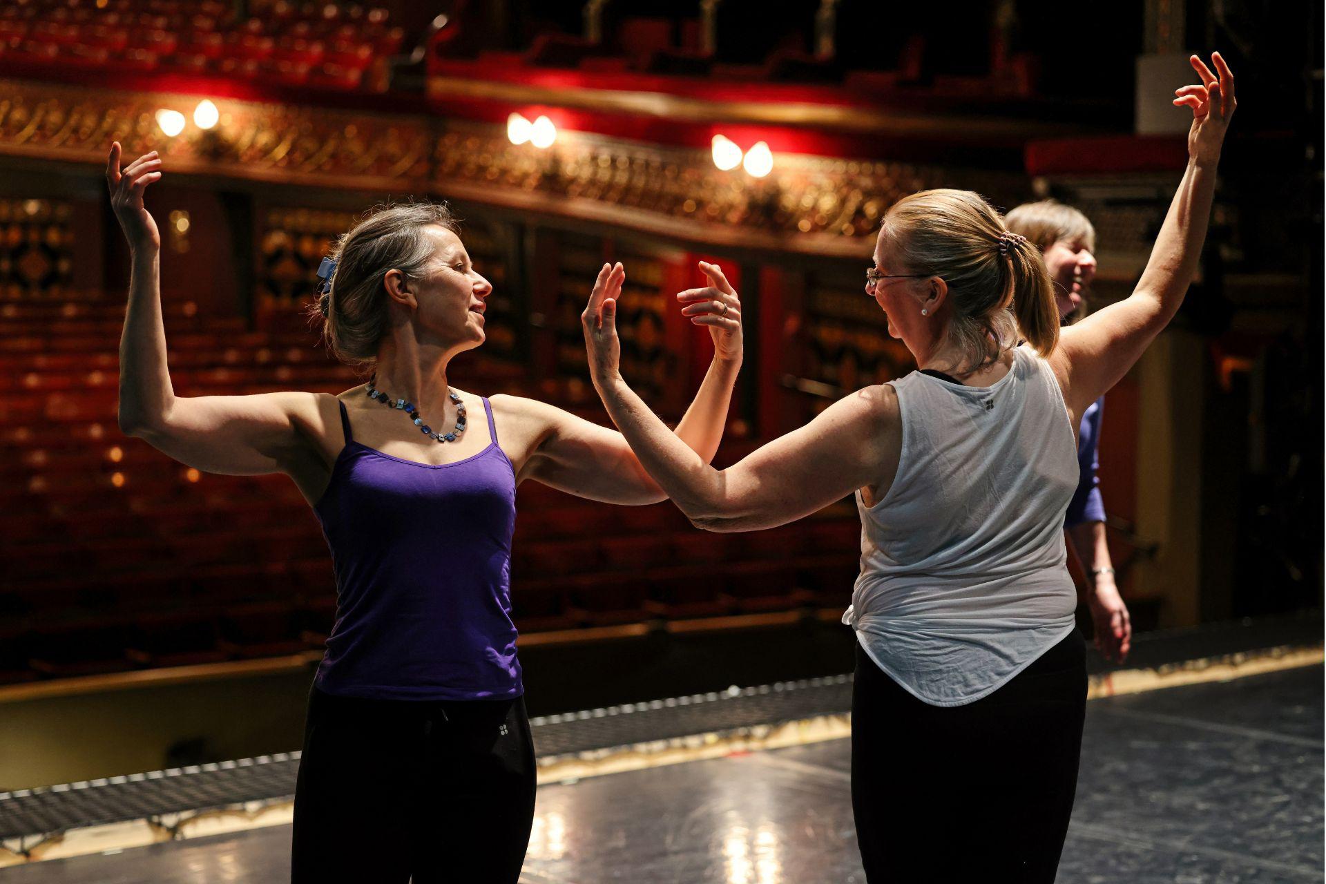 Two older dancers rehearse moves on a stage in front of an empty audience space. They stand side-by-side facing opposite directions. Their raise both their arms, nearly touching elbows whilst looking at each other.