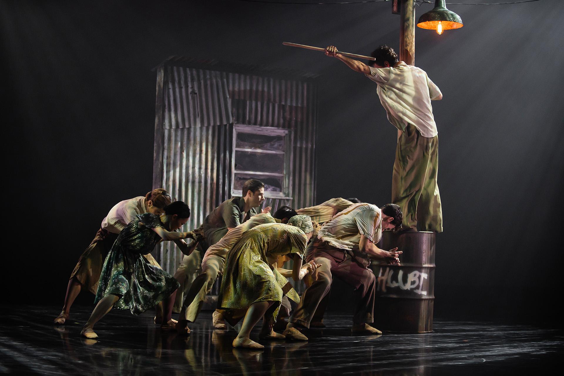 A group are each leaning low, as if burdened, another stands on a metal drum shouting at them