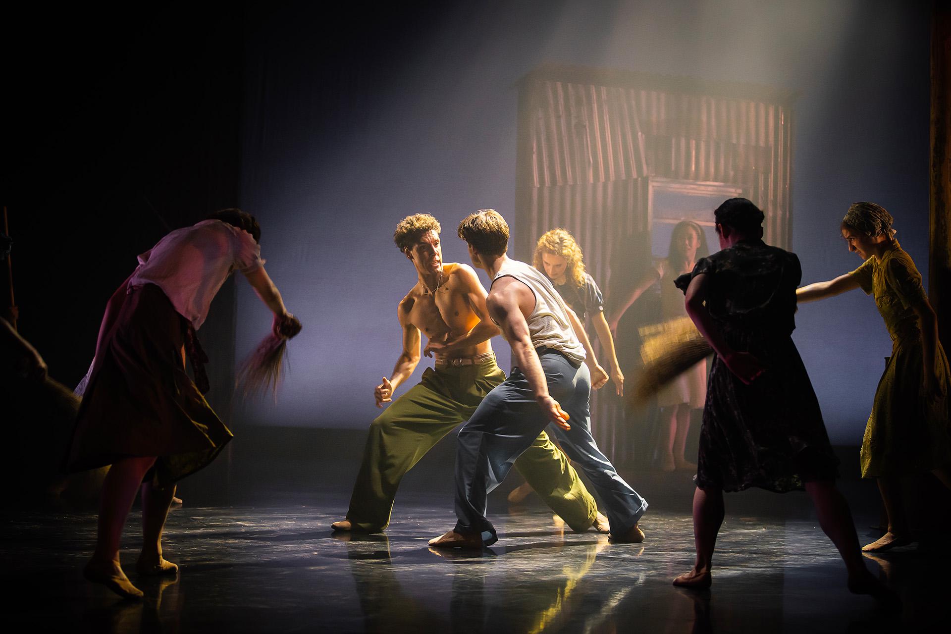 Two dancers, one bear chested in green trousers, the other in blue trousers with a white vest, fighting, one turned slightly and leaning back with an angry expression about to punch the other