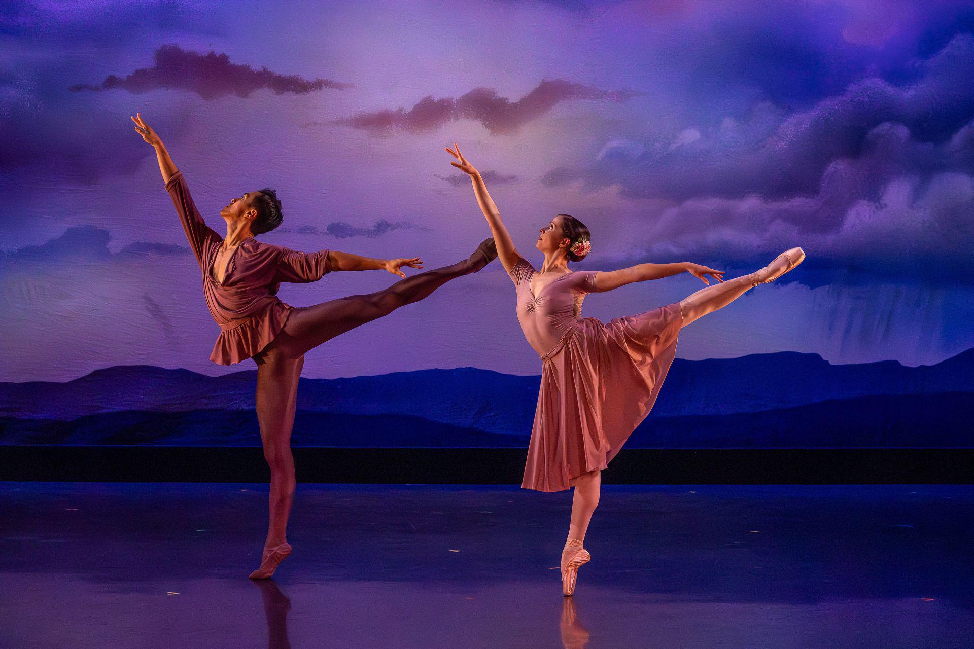 Two dancers lift their leg in the air and reach an arm upwards in unison