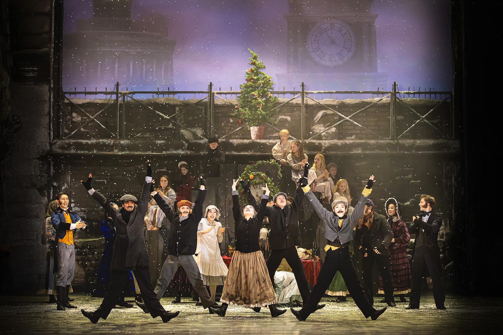 Townspeople dance with their arms in the air and on their heels as snow falls.