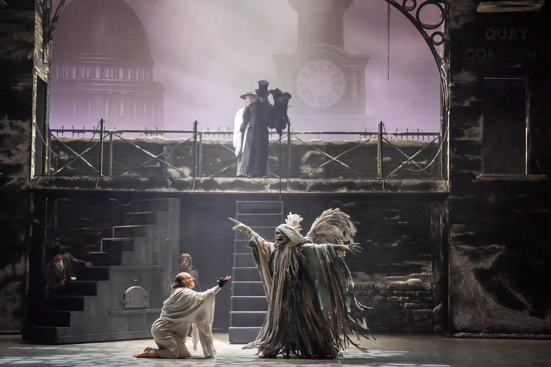 A Christmas Carol, 2024, production, dancer Andrew Tomlinson and Jonathan Hanks, Photo Tristram Kenton Scrooge begs at the feet of the Ghost of Christmas Yet to Come. A figure dressed in black and a top hat stands at the top of a set of stairs behind them.