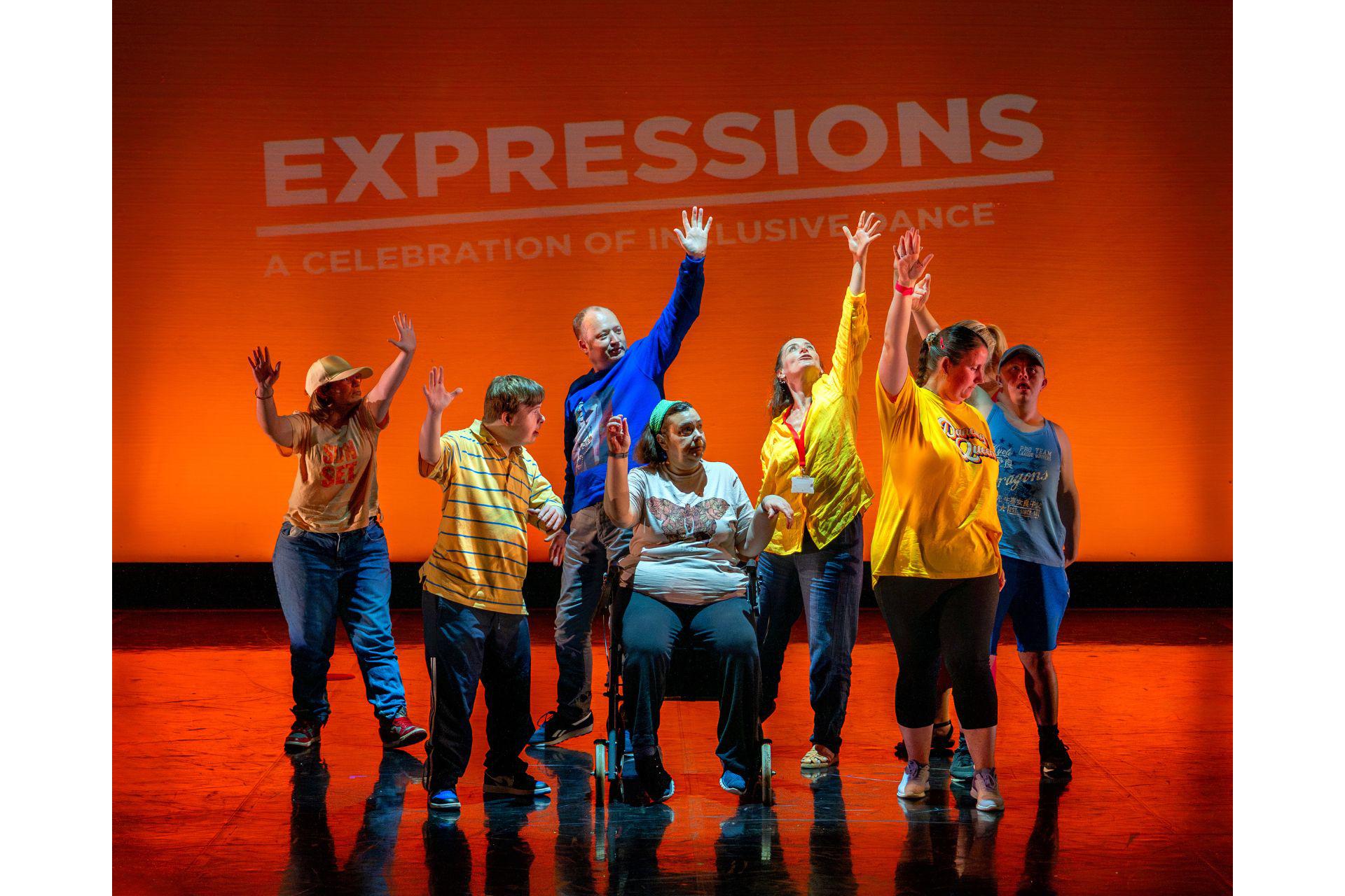 A group of dancers on an orange stage raise their hands.