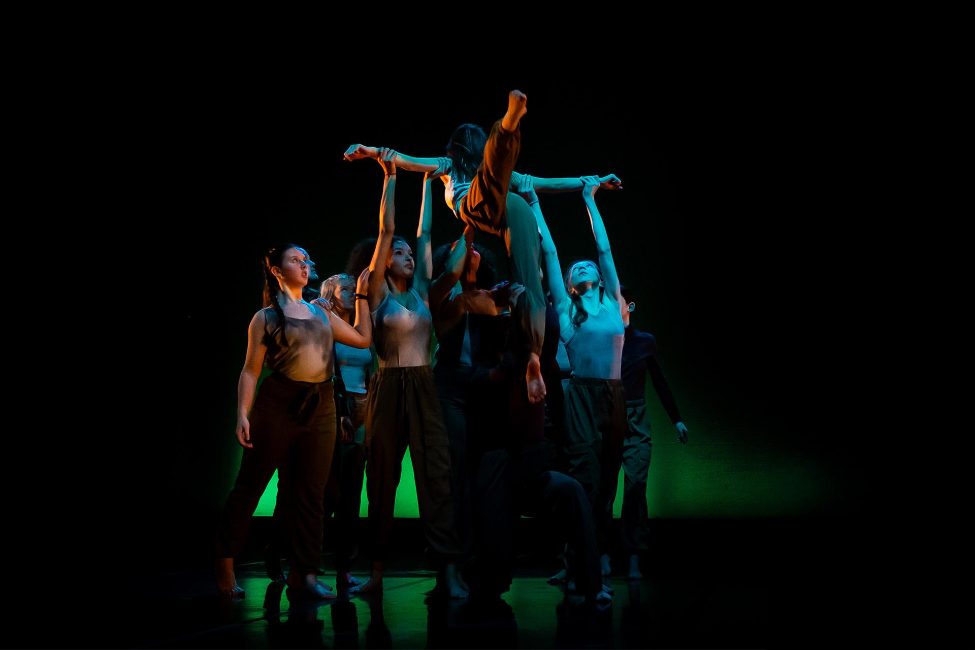 Against a black backdrop, many dancers hold up one with their arms apart, one leg arabesque and the other pointing to the ground
