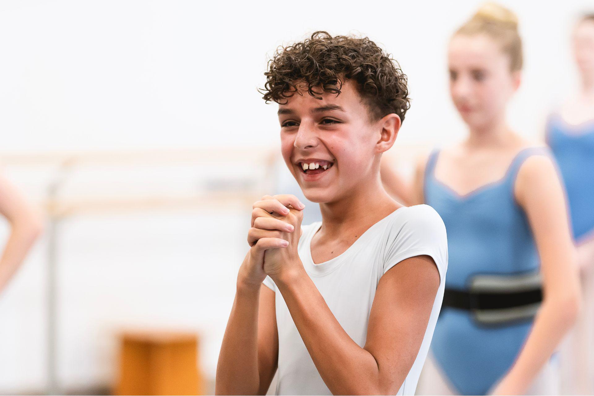 A child in a ballet studio interlocks his hands and laughs.