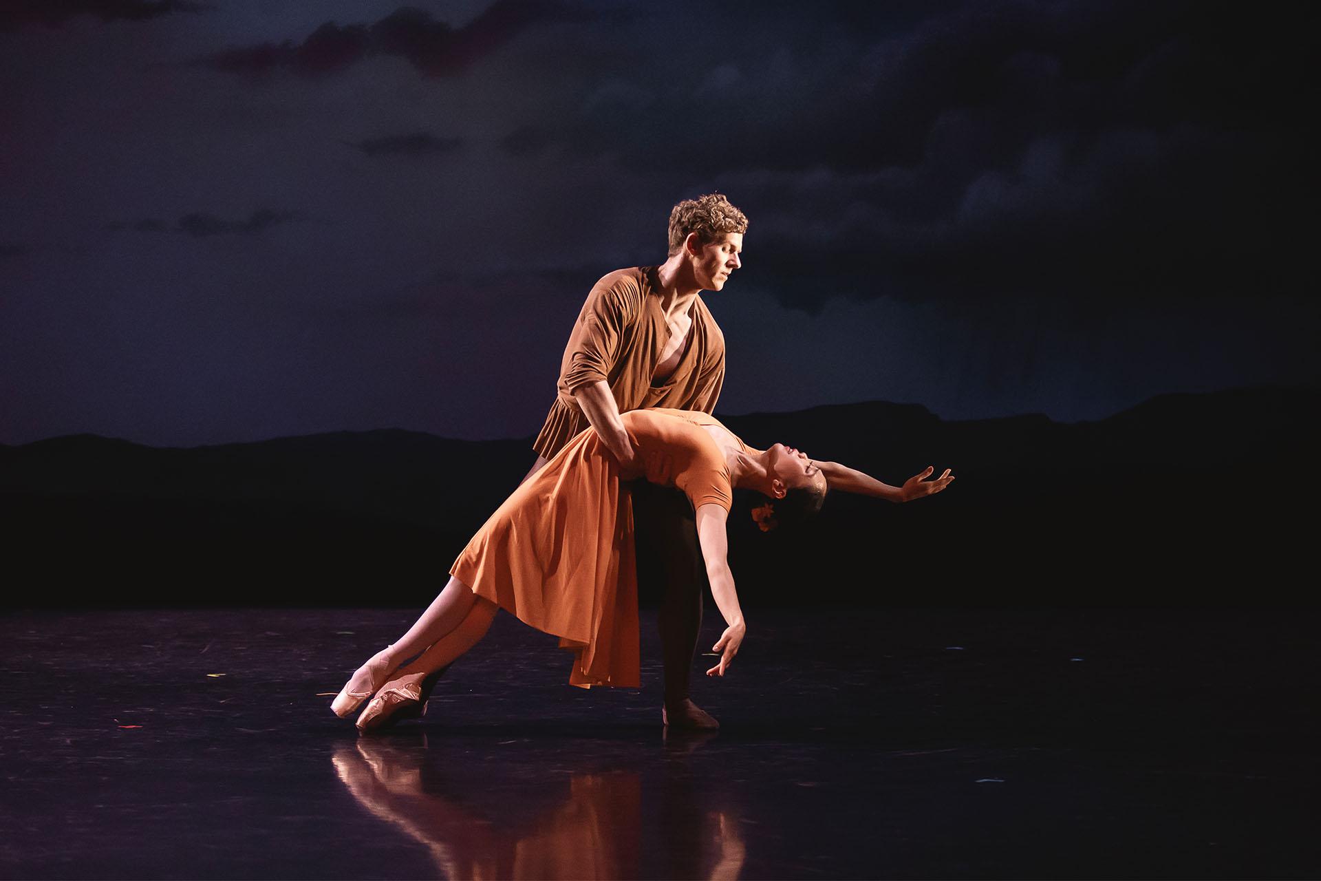 Dancer in an orange dress leans back, she is held by a man in brown with his arms around her waist