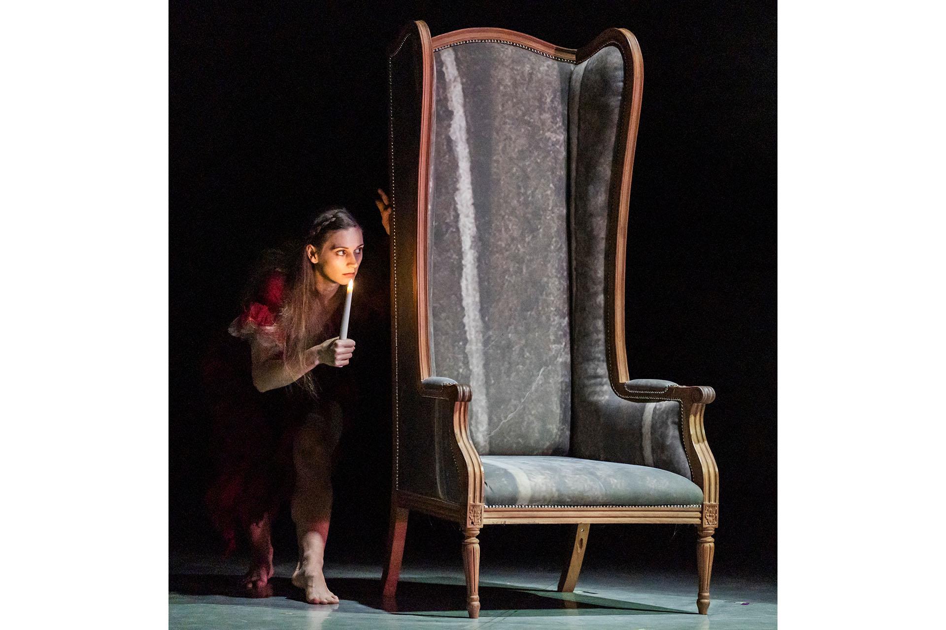 Jane Eyre, production, 2025, dancer Amber Lewis. Photo Tristram Kenton Lit by the flame of a candle, a mysterious figure, partly hidden by a large-backed chair