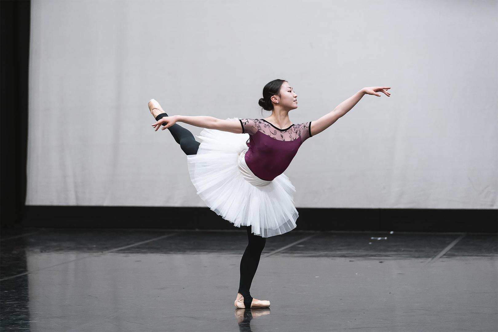 A dancer stood on one leg with the other raised and arm extended. The dancer is wearing a dark purple leotard, black leggings and a white tutu