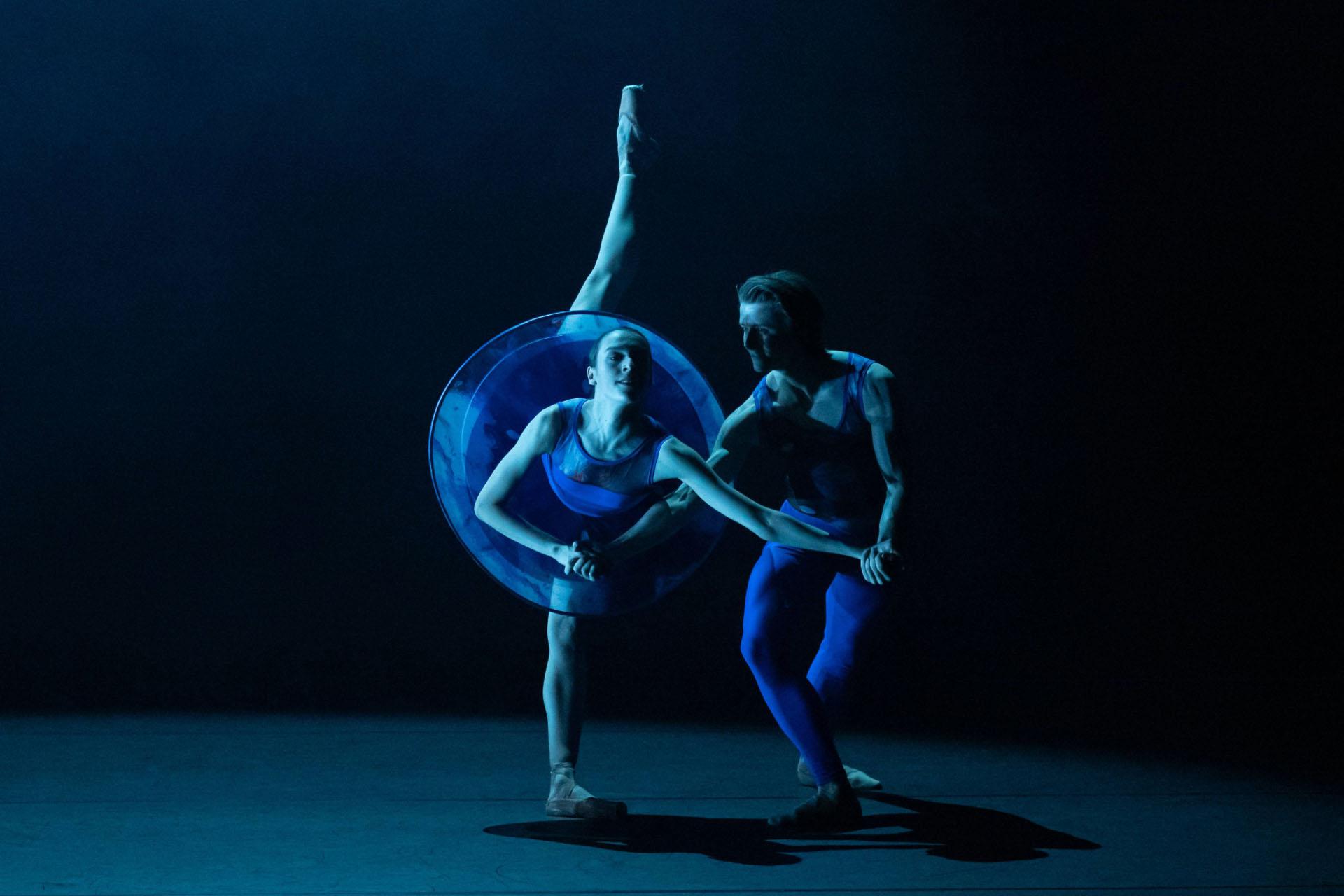 Bathed in blue light, two dancers pose, one dressed in a tutu, the other wearing a leotard