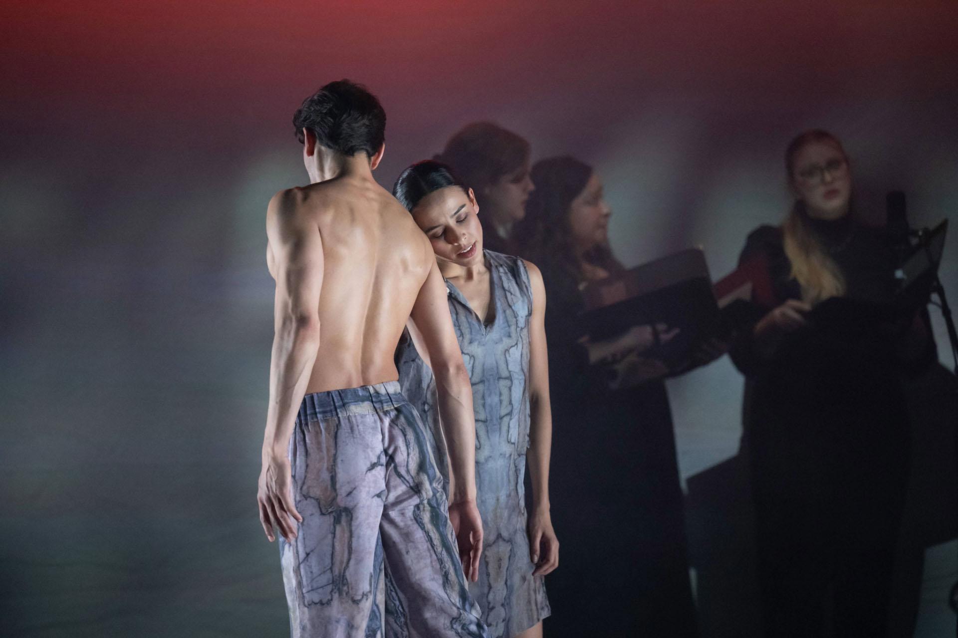 Two dancers facing opposite directions lean against each other, eyes closed, wearing grey clothing