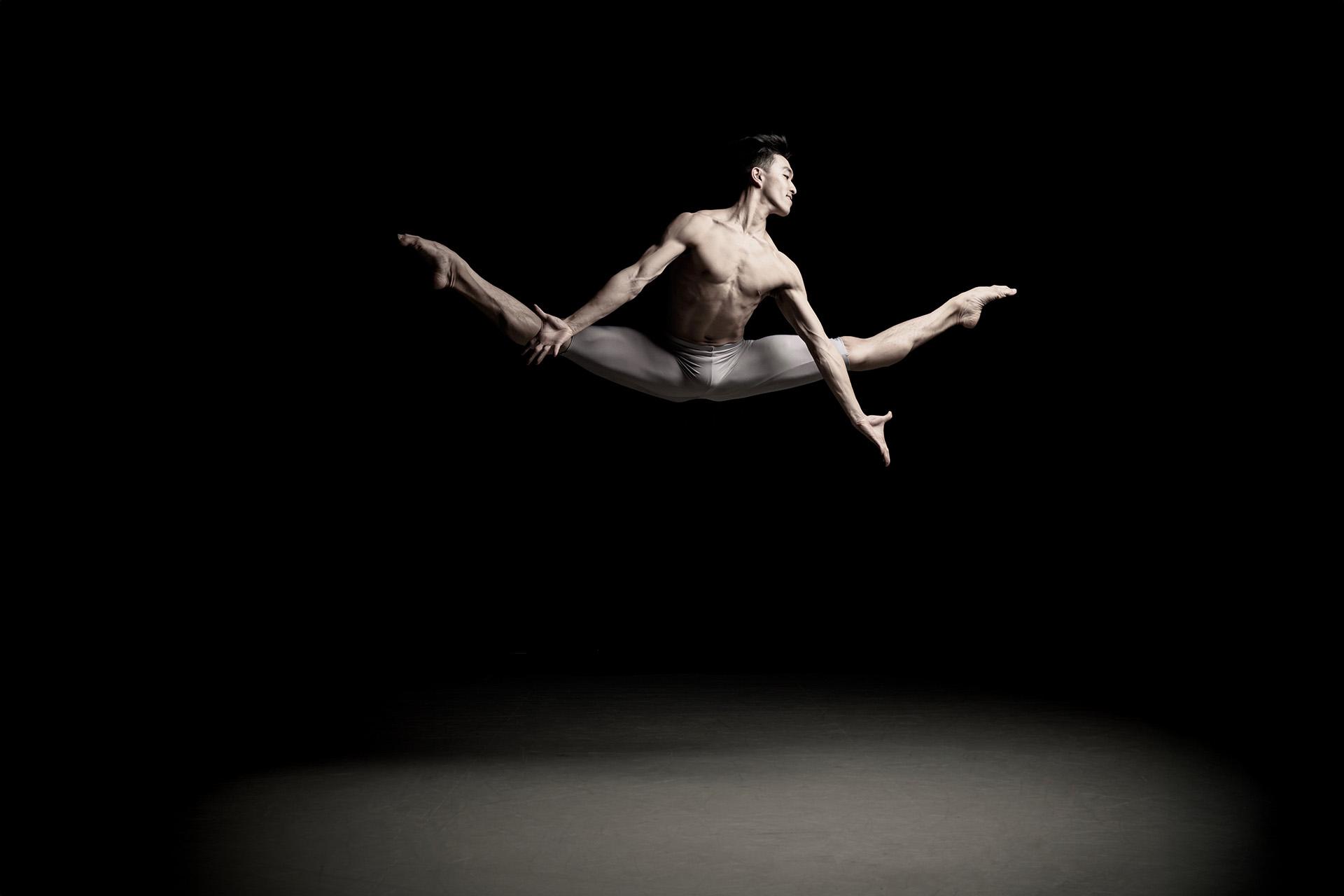 Male dancer, only wearing white leggings, mid jump, legs split, arms reaching down
