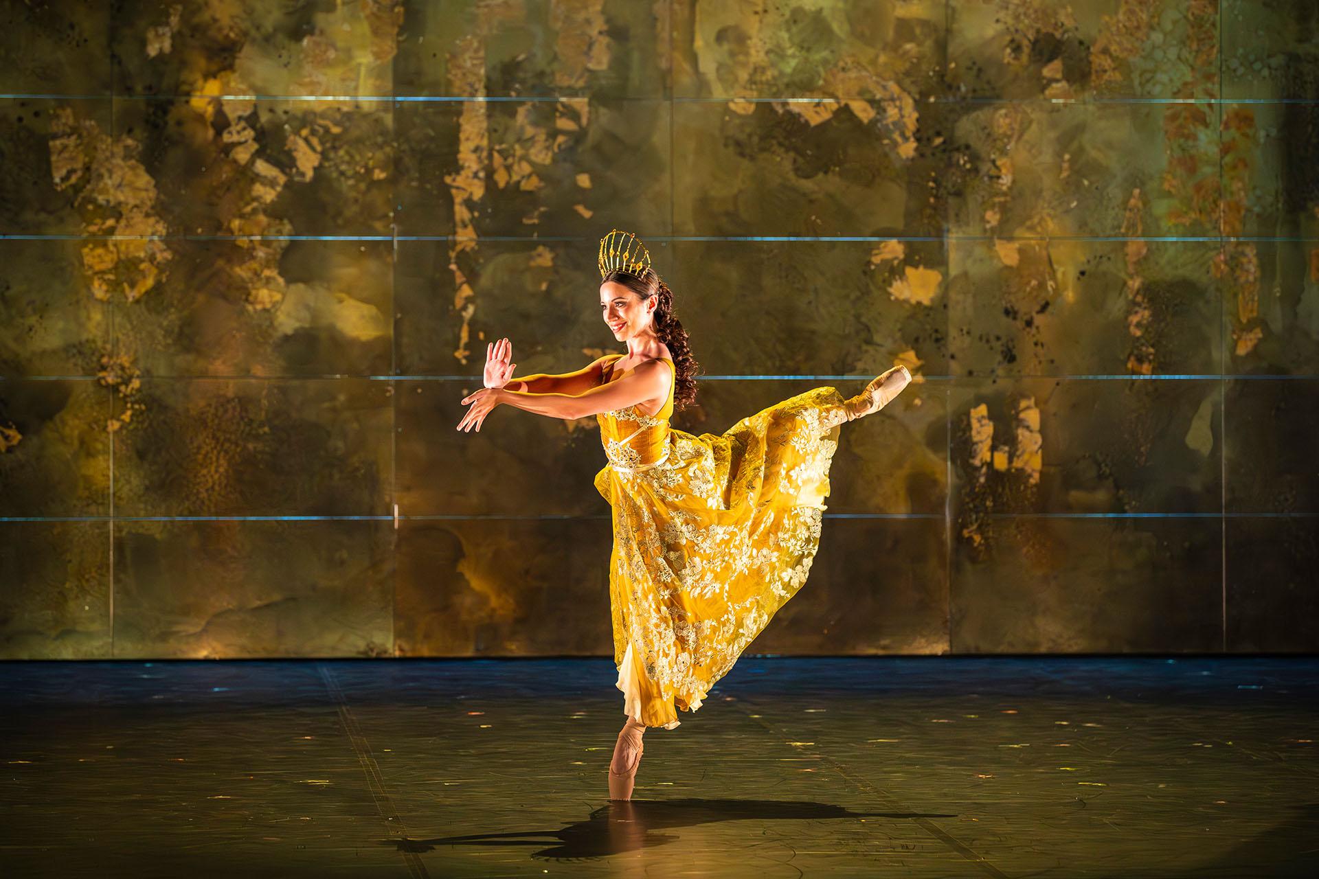 Merlin, 2025, production, dancer Alessanda Bramante, photo Emily Nuttall Dancer in a yellow dress wearing a golden tiara stands arabesque