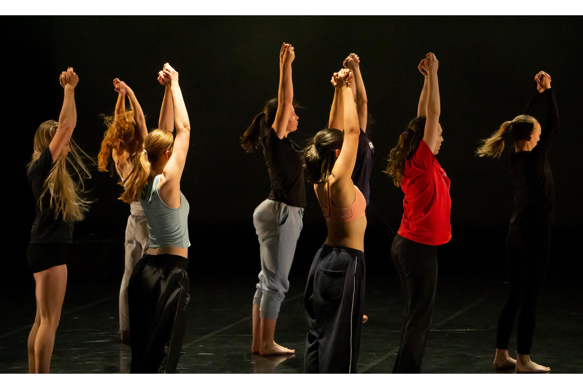 Troup of dancers standing side on to the camera, each with their hands joined above their heads, arms straight