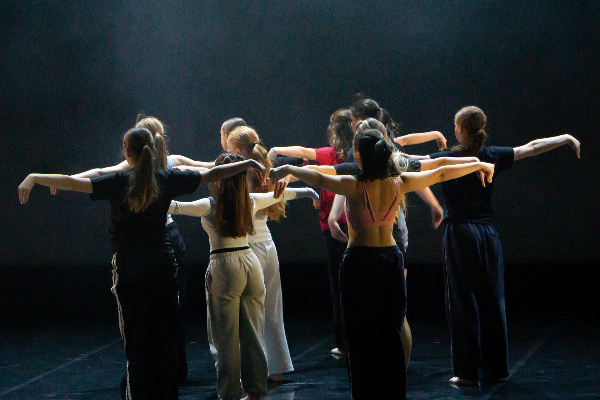 Troop of dancers turned away from the viewer, each standing with their arms straight out level with their shoulders