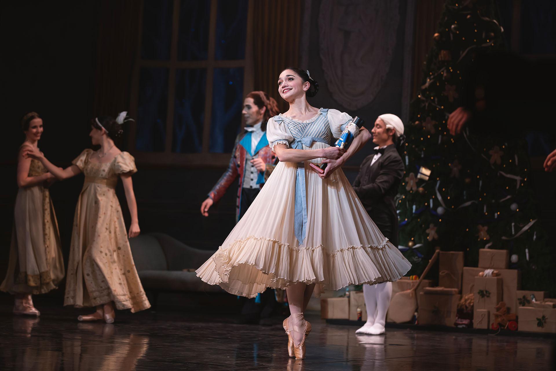 A dancer in a white and blue dresses stand on pointe and smiles while holding a wooden nutcracker doll in her arms.