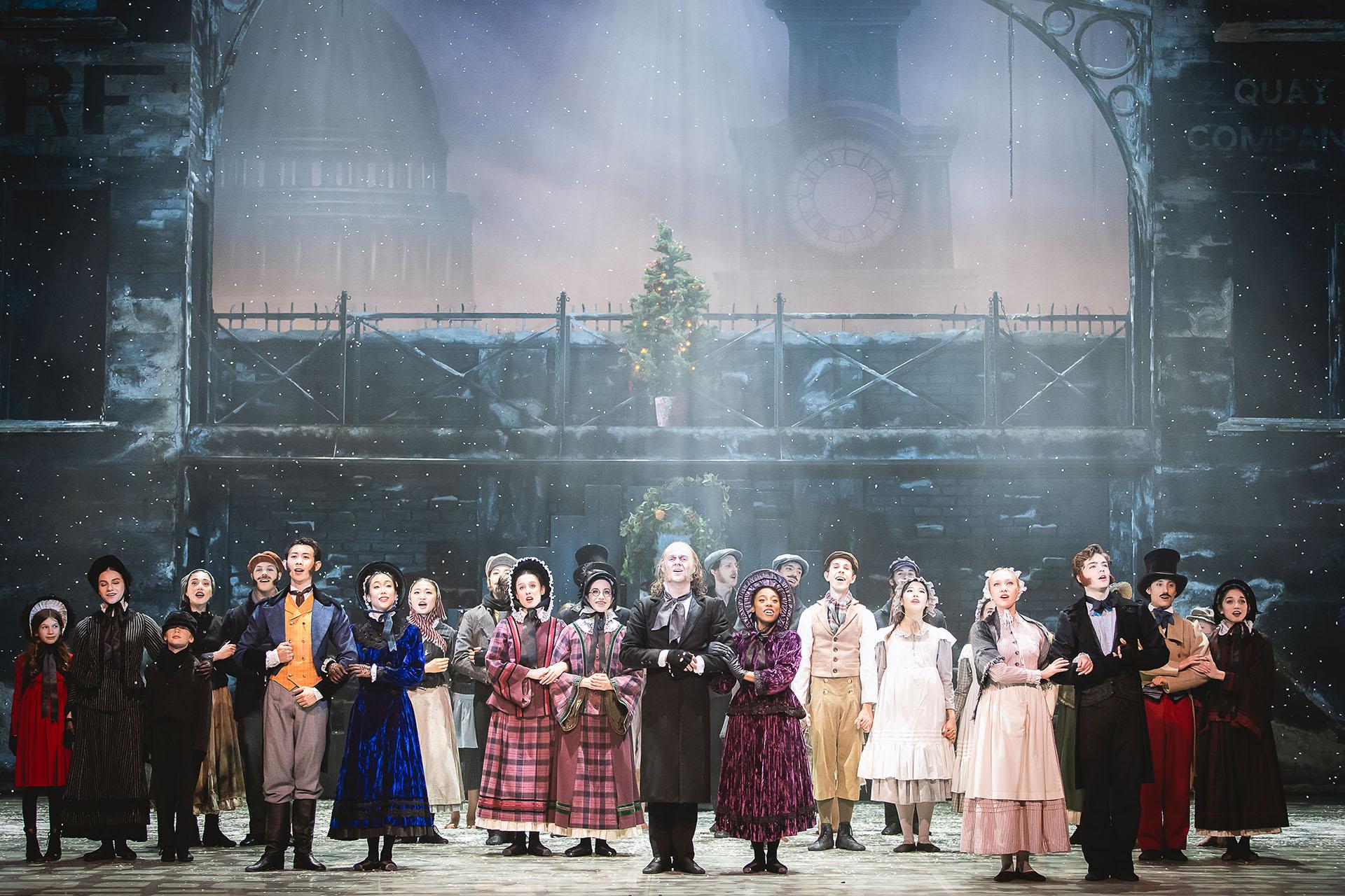 The cast of A Christmas Carol stand on set looking out with Scrooge at the centre and the set of Victorian London visible behind them