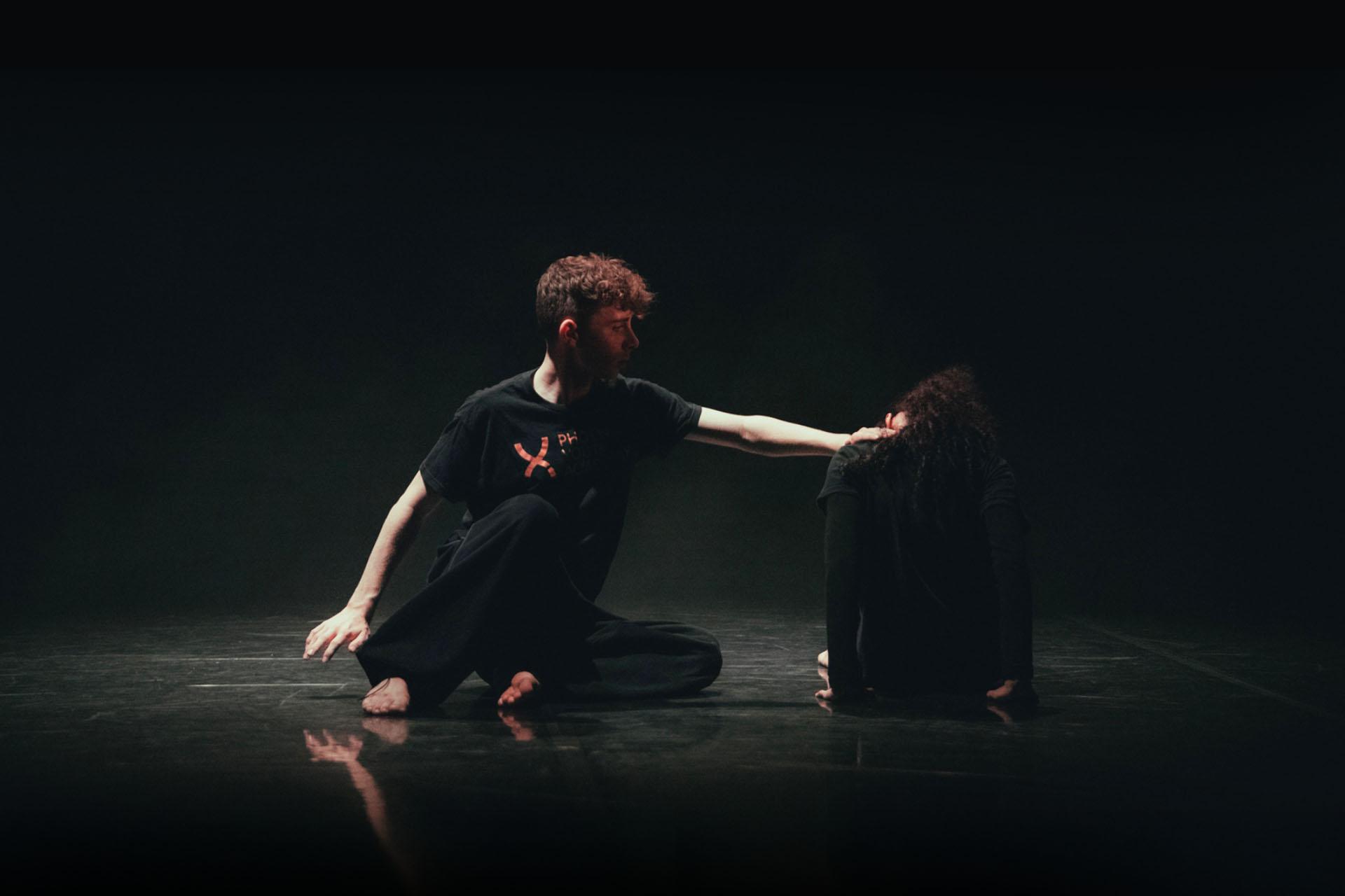 Dancers on the dark floor , lit sparingly from above, one reaching out to the other who has their back to us and head down