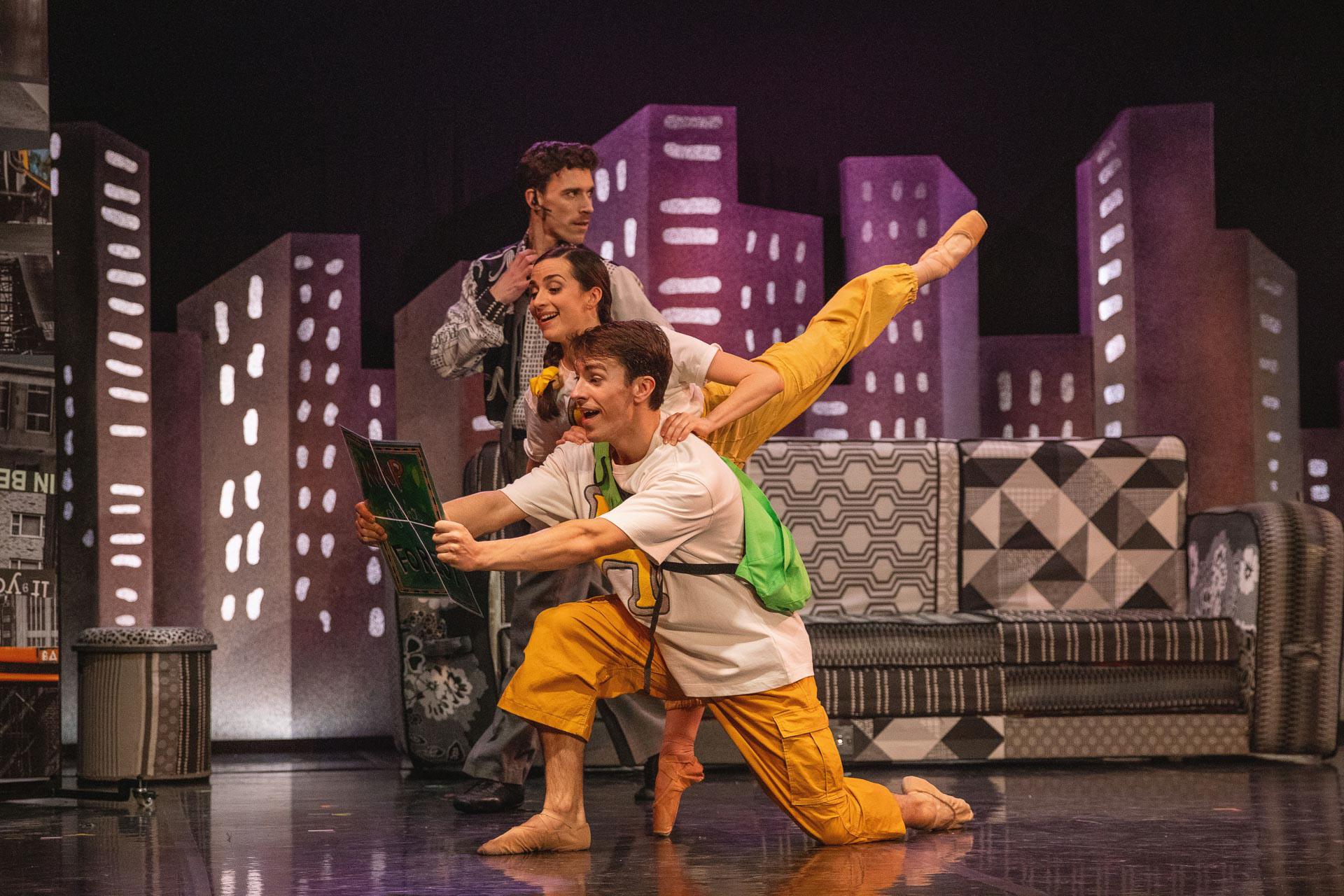 Two dancers look at a map, one over the shoulder of another with leg raised high