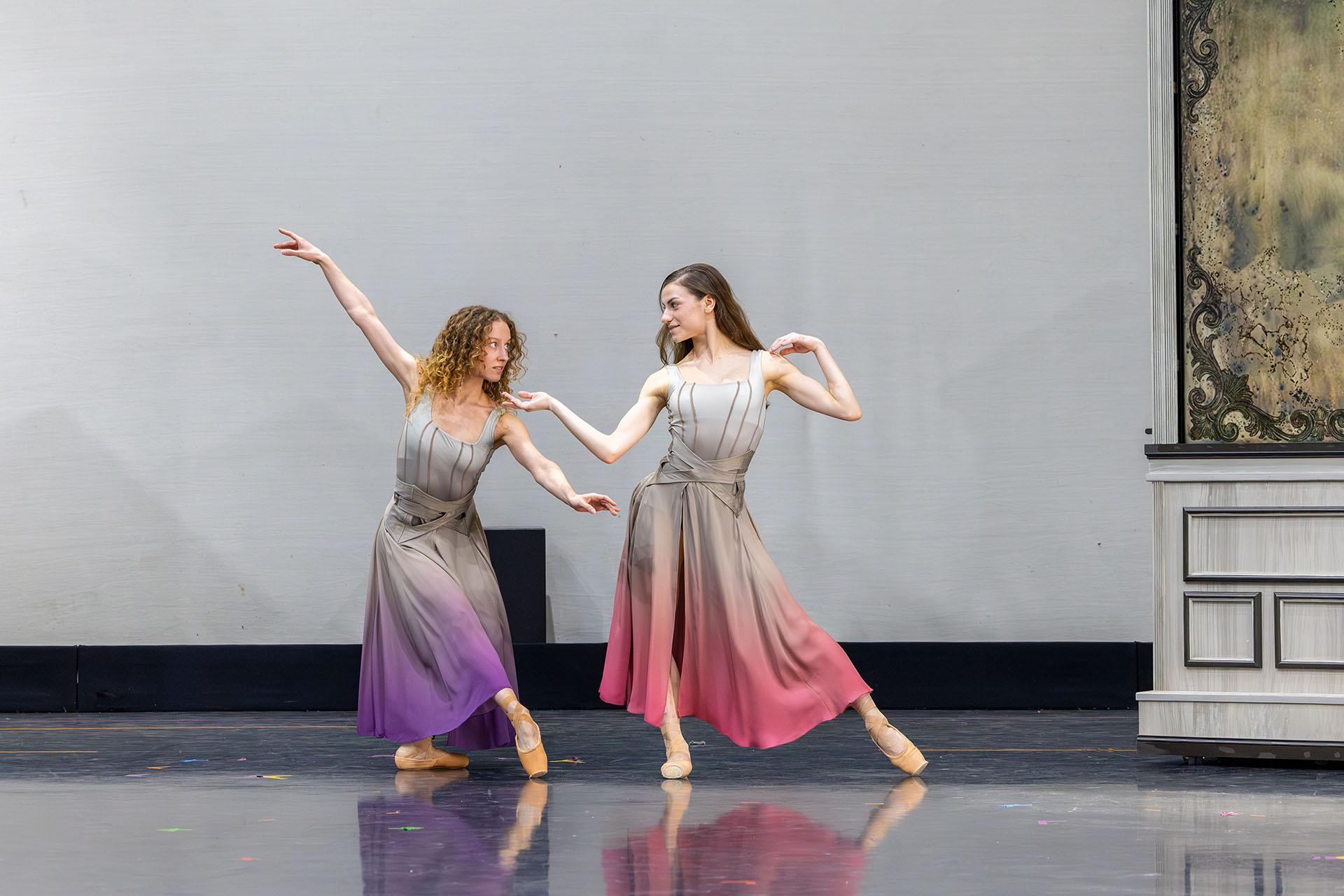 Two women in grey dresses, one with lilac trim, the other with pink trim, dance while sharing a look
