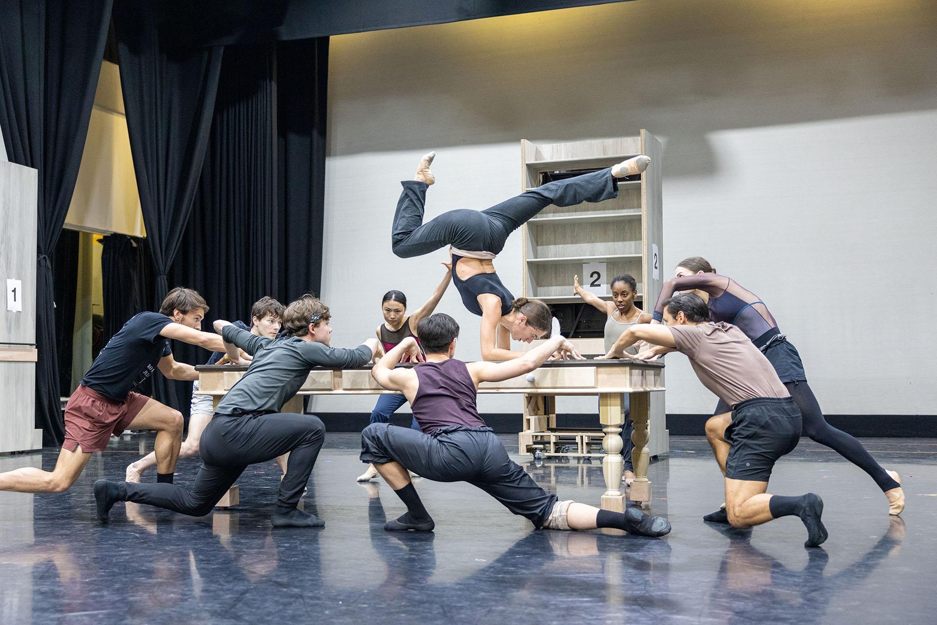 A woman in dark clothing hand-hands on a table, her legs split, around the table are other dancers crouching down looking at her