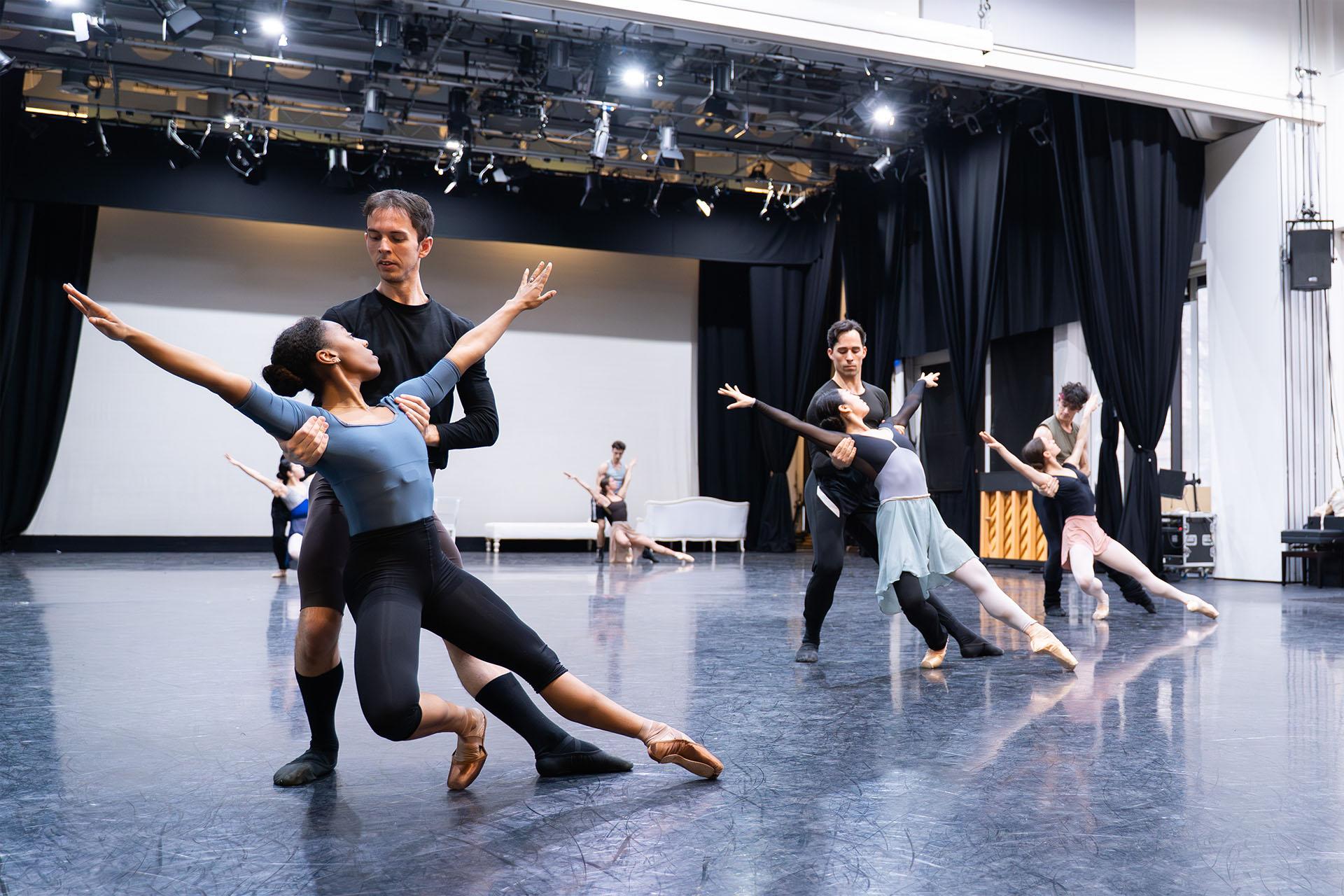 Three pairs of dancers in the foreground, two in the background, each in the same pose - a male dancer behind a female holding her under her arms, her right leg beny, but toes of both feet on the ground, and she's looking up at him