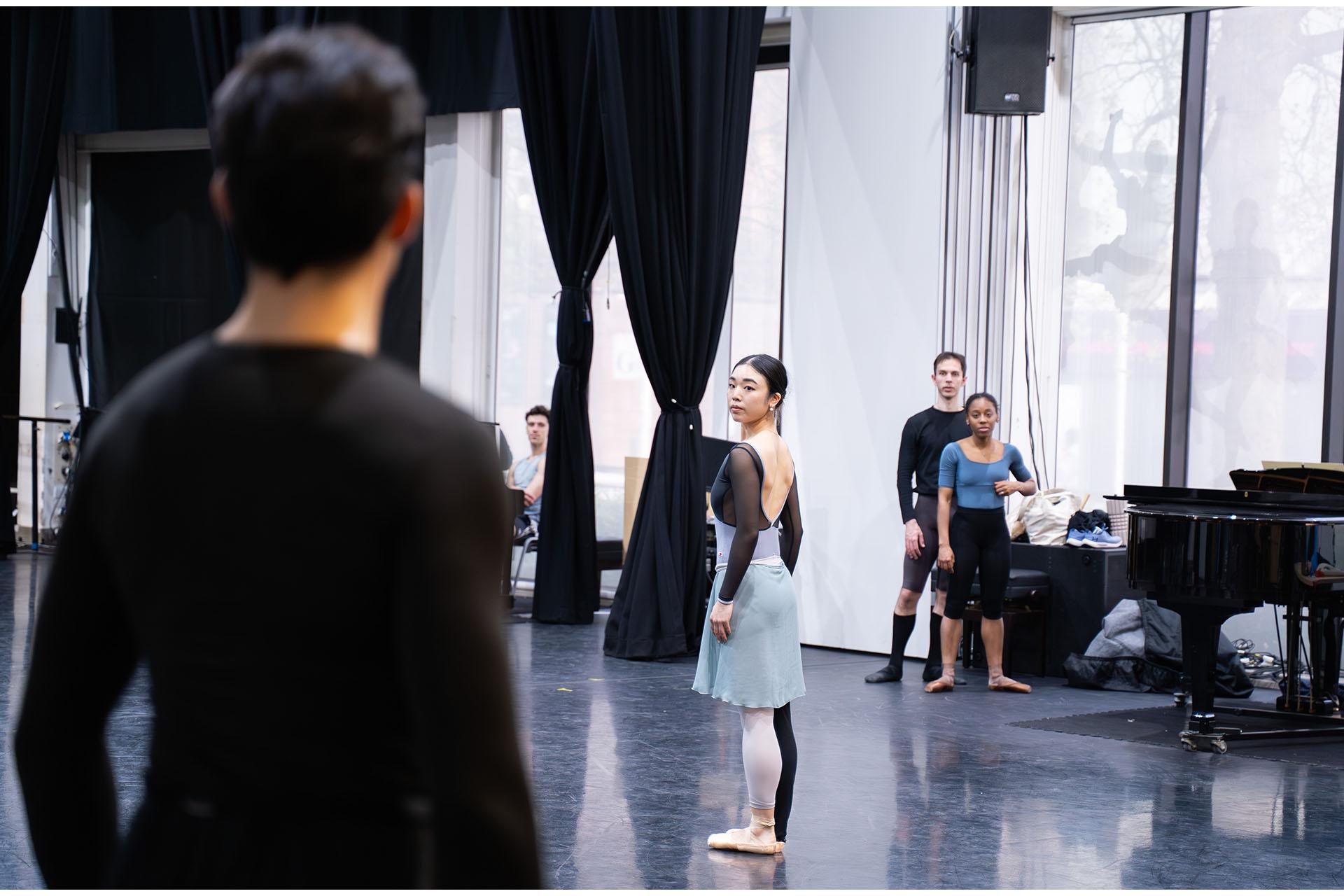 Dancer with tied back dark hair stands straight, body turned from the camera, looking over her shoulder at another dancer, blurred next to the camera