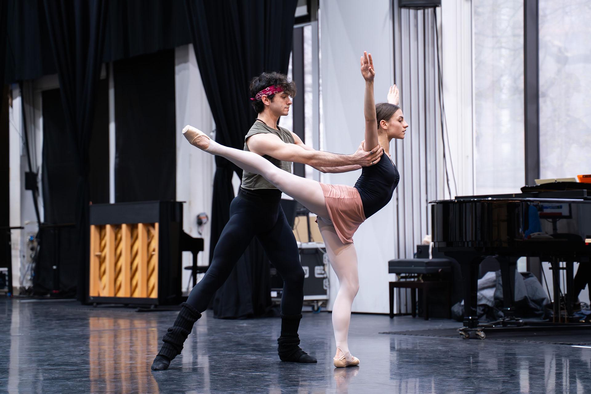 Dancer stands on one foot, herother leg exteded behind her, arms raised above her head, another dancer standing behind her holding her steady under her upper arms