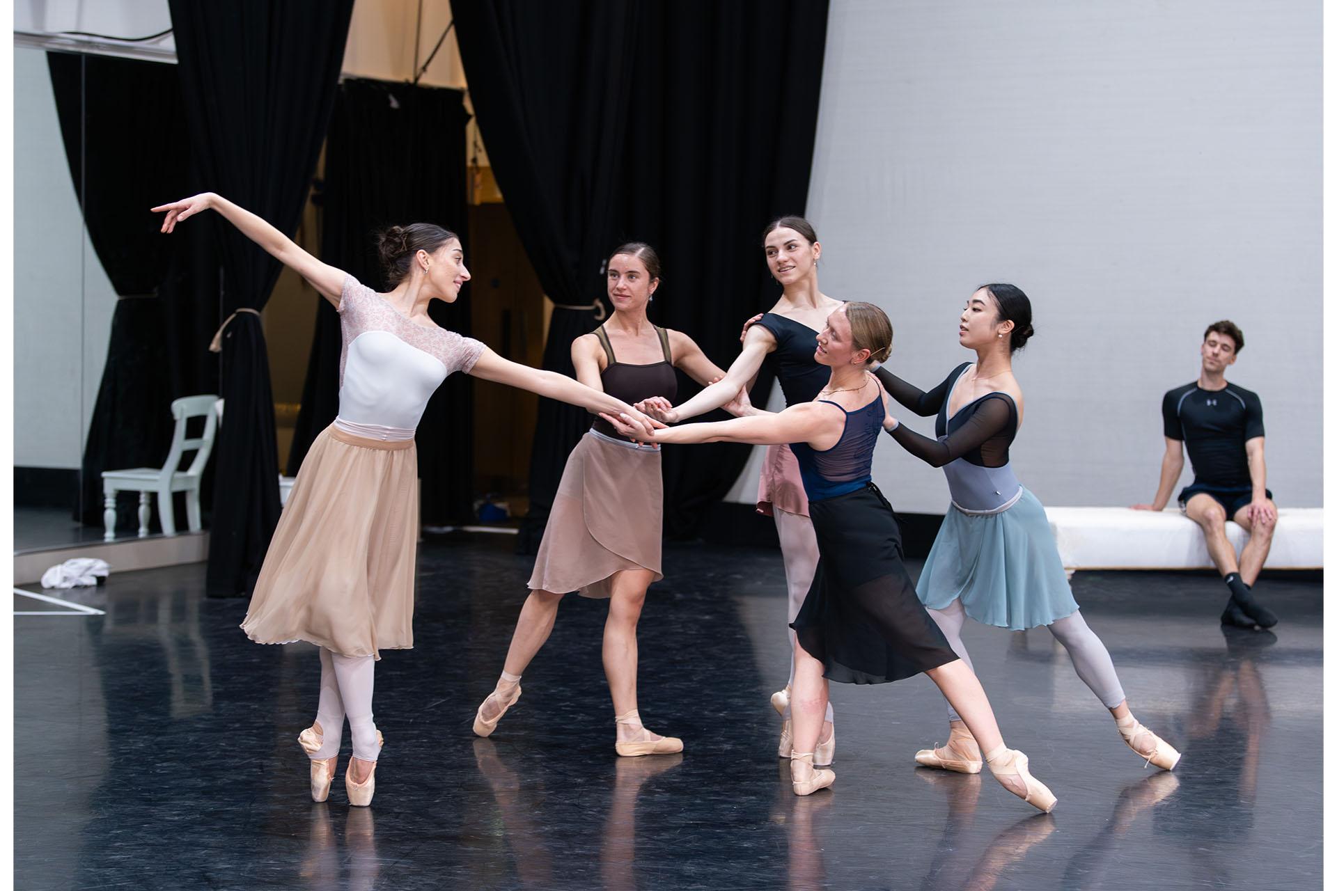 Five dancers in the rehearsal room in a circle all touching hands in the centre, one en pointe