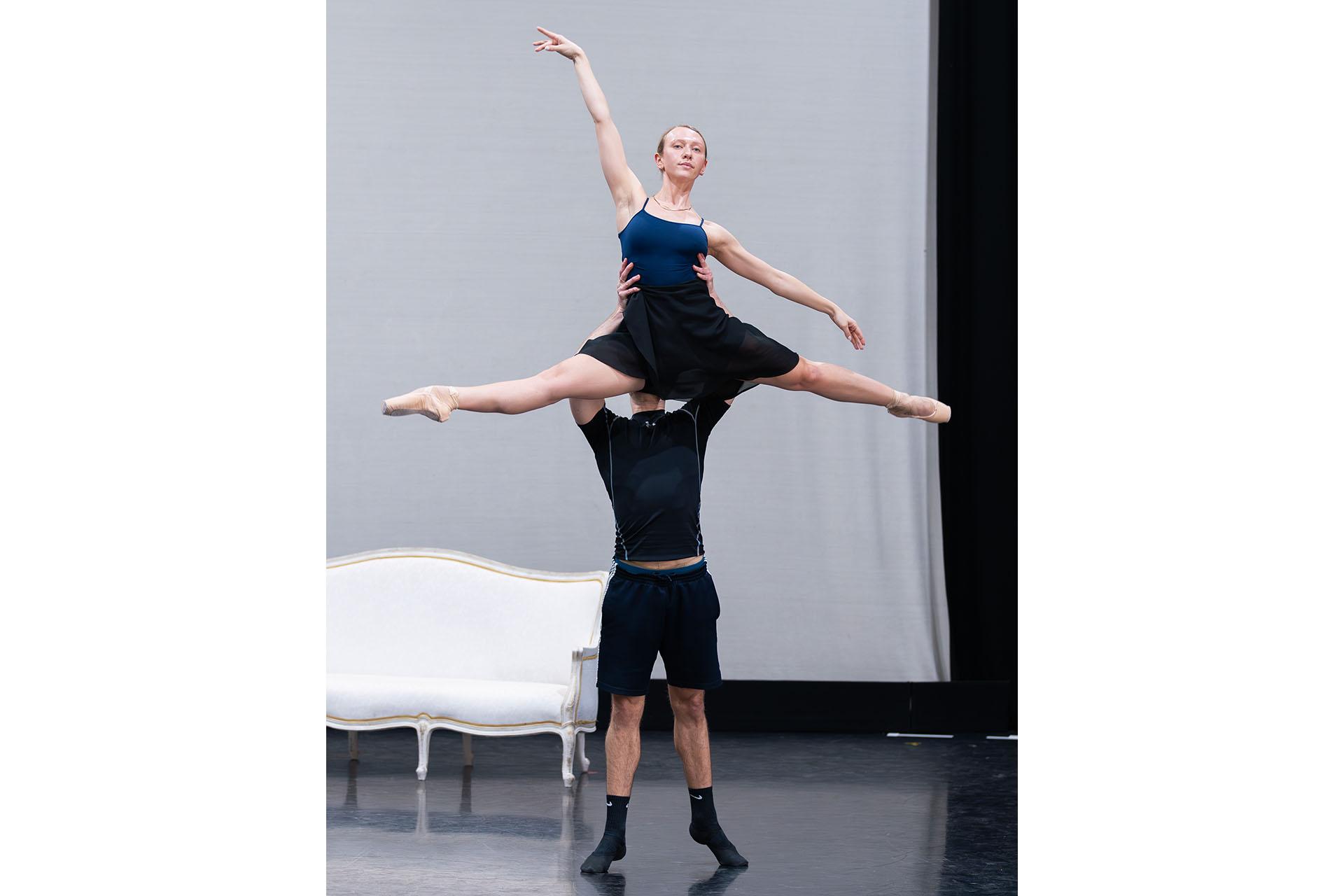 In rehearsals a dancer is lifted by the waist, one arm straight above her head, legs straight forward and back