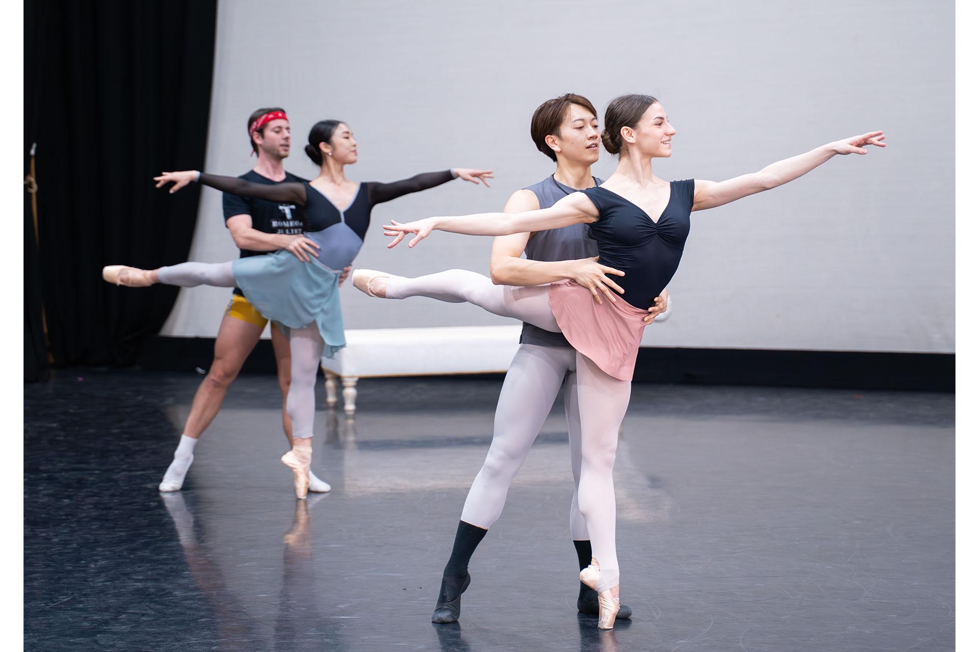 A pair of dancers in rehearsals, one stood behind the other supporting her balance by holding her waist, the woman on one foot, en pointe, leg straight behind her, another couple further in the distance in the same pose