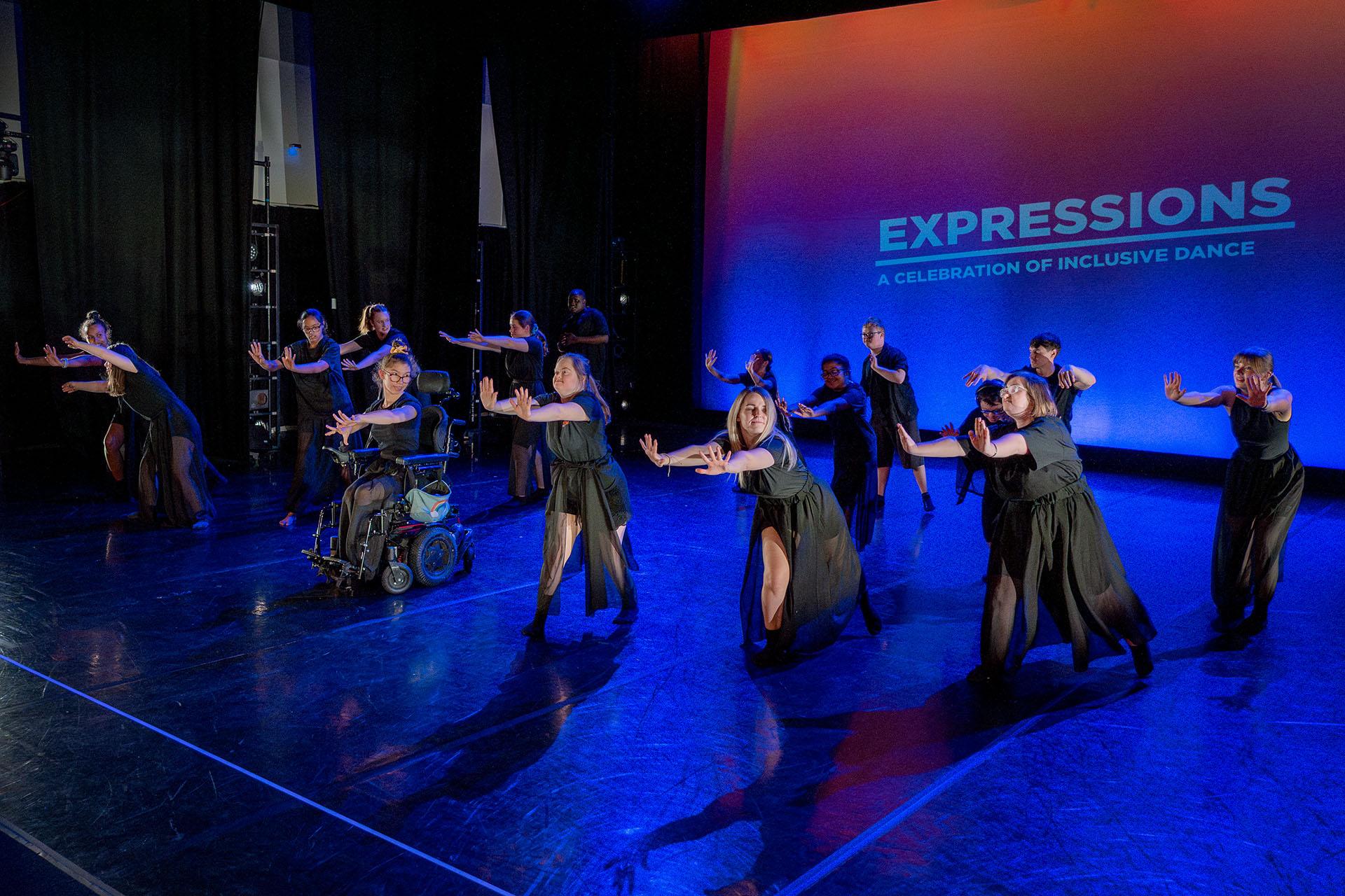 A group of dancers wearing dark grey, each leaning forward with their arms help out, palms forward to the audience