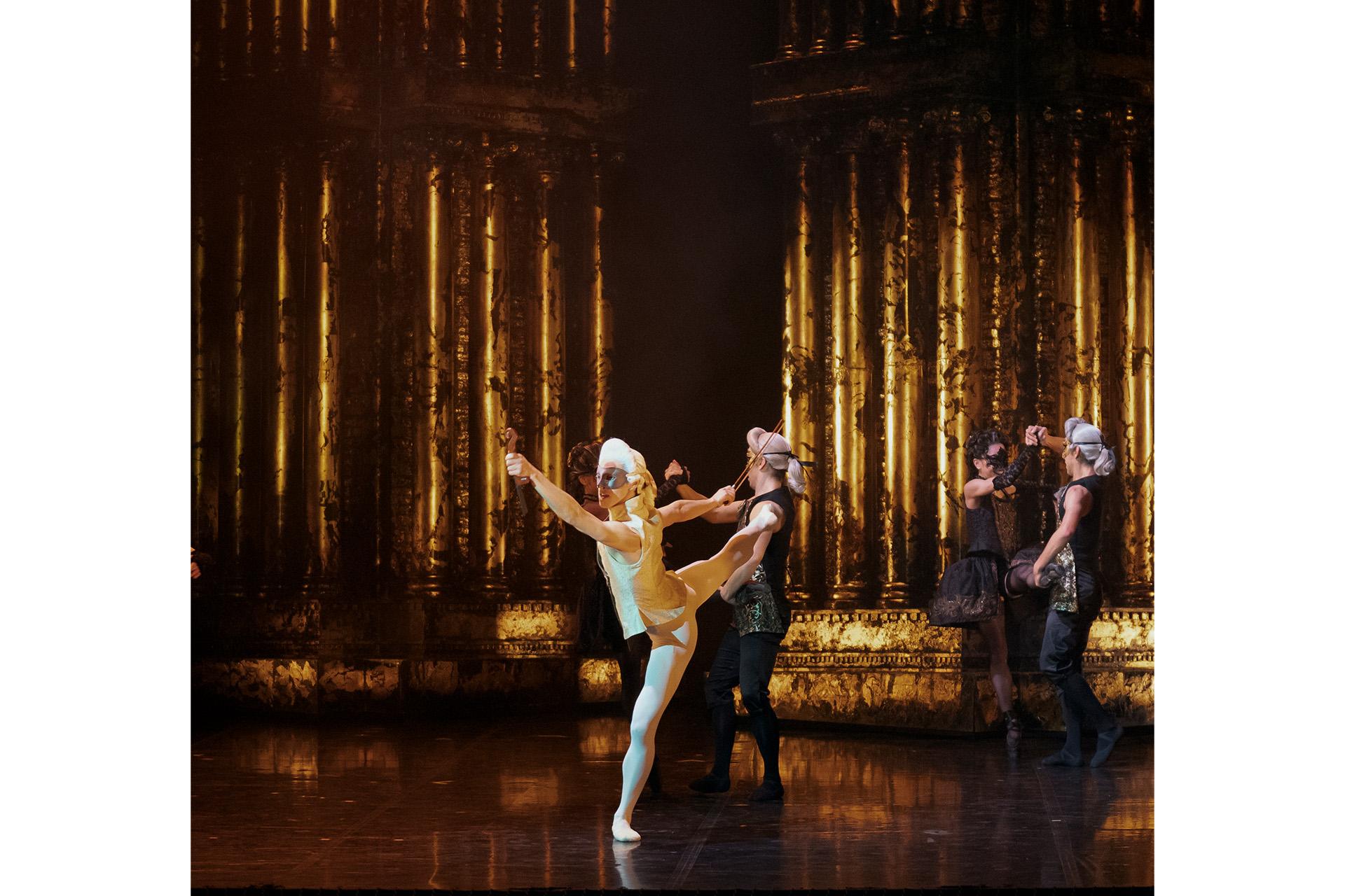 Dancer in white wig and dark mask holding a conductor's baton and standing arabesque