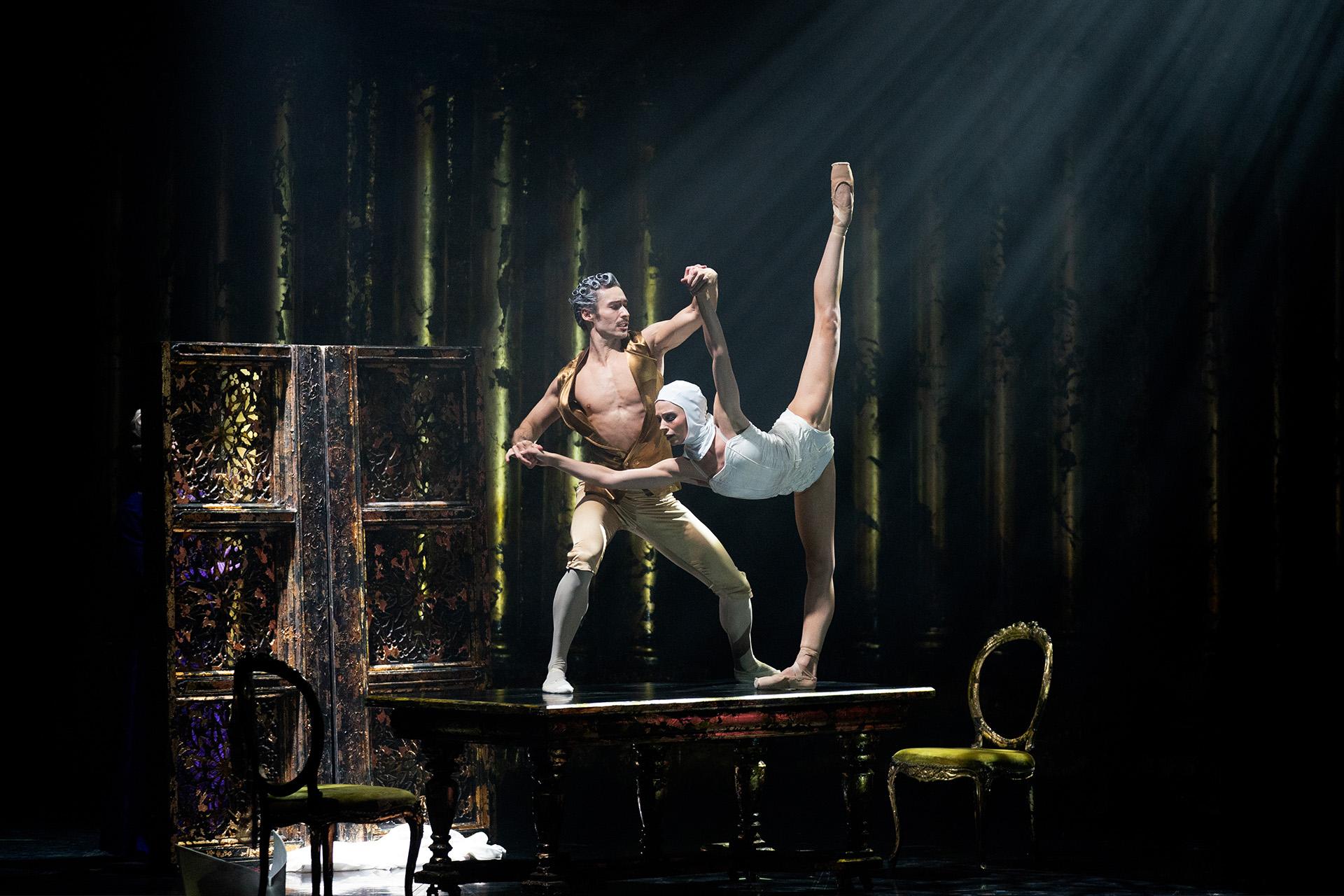 Two dancers on a table. One male and shirtless wearing golden britches, the other standing straight legged on one foot the other leg high behind her and body, in white, leaning forward and supported by the man