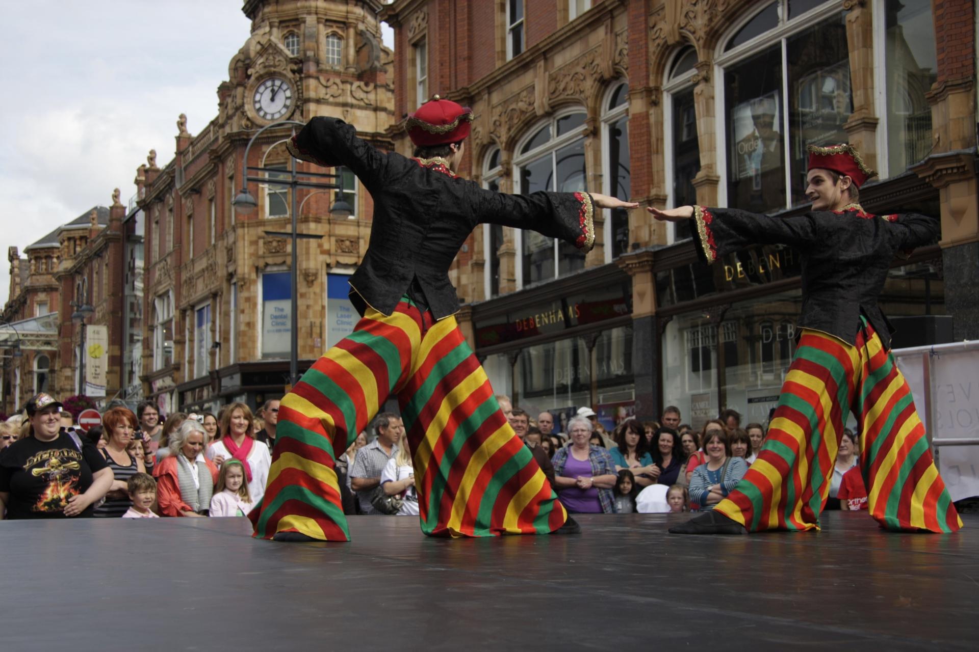 Two dancers in colourful stripped trousers and black tops pose while dancing on Briggate in Leeds. Spectators watch in the back.