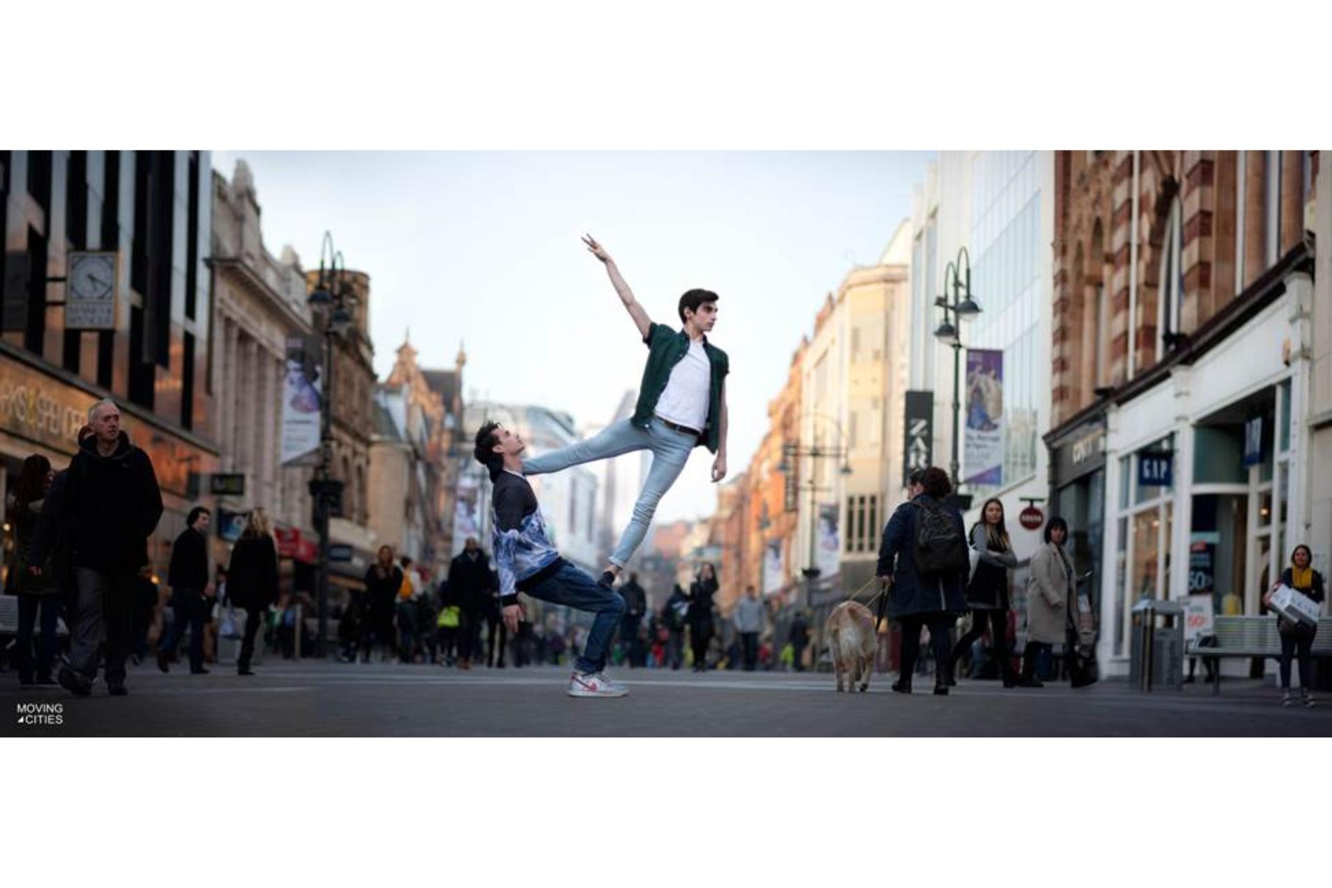 Two dancers strike an acrobatic pose, with one holding a sitting position while the other balances on top, in a Leeds city landscape