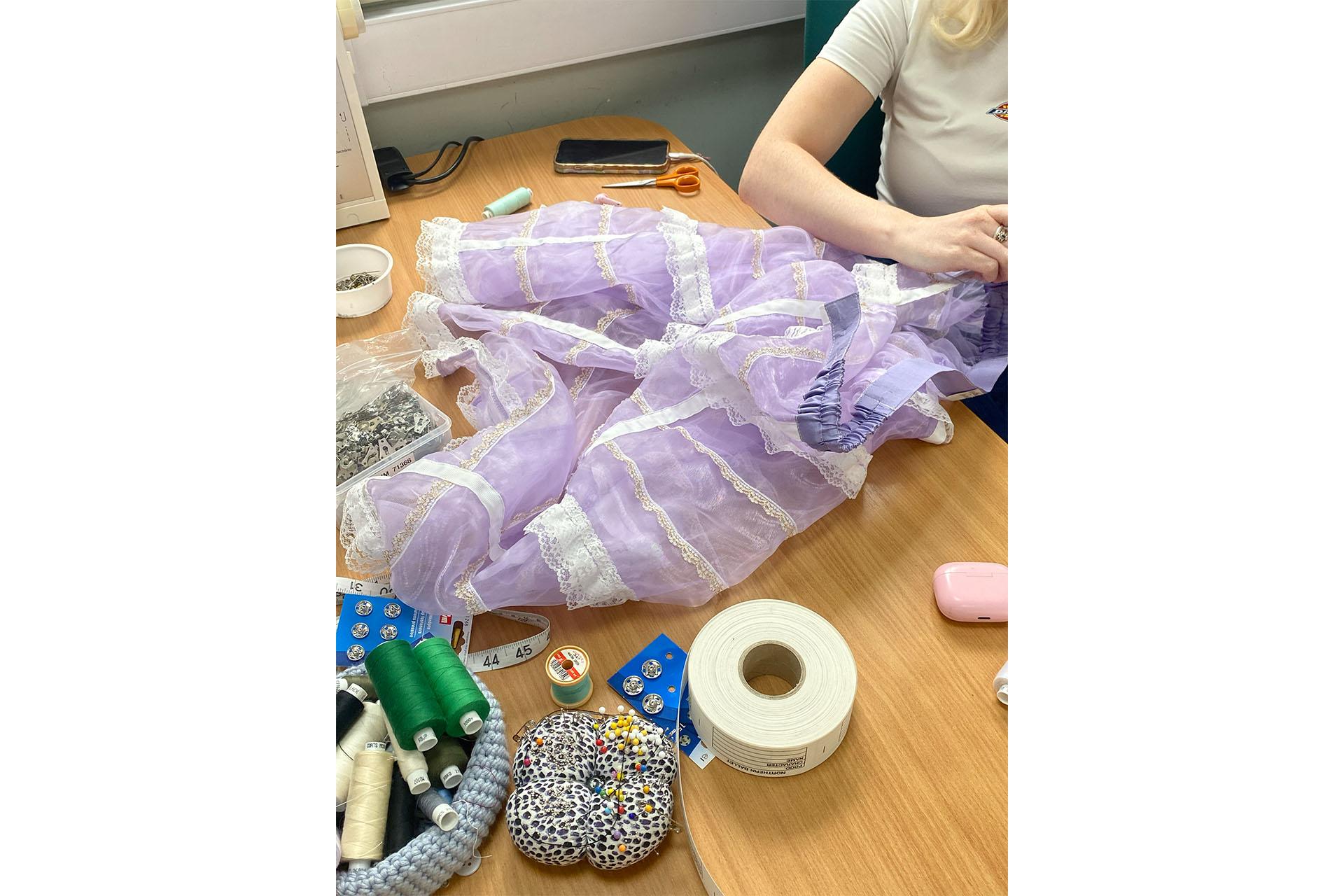 A lilac dress having white lace sewn on at a desk which is full of thread, scissors, measuring tape and more accruement from Wardrobe