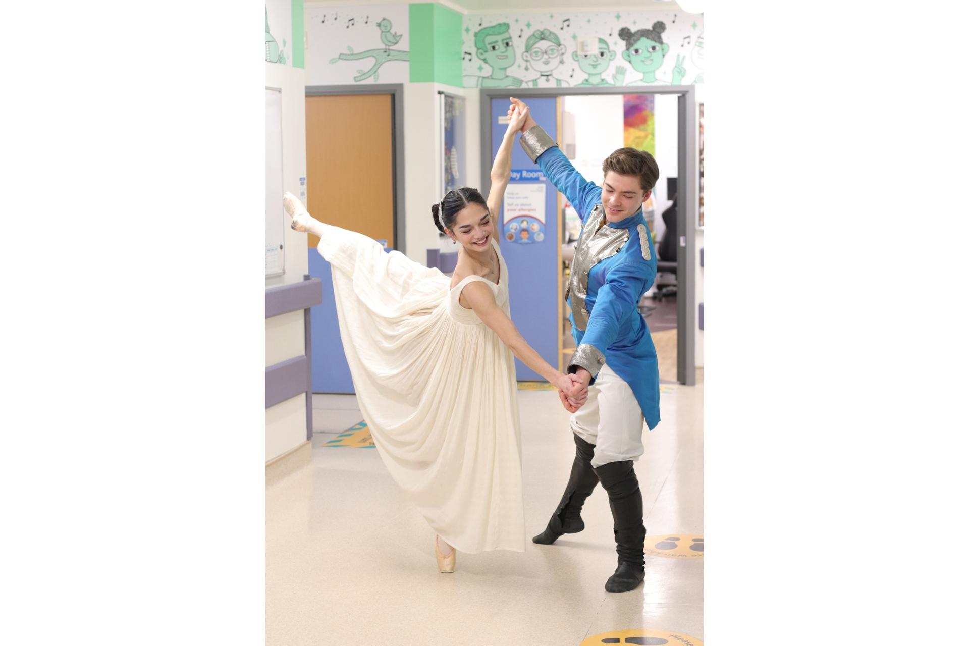 Two dancers, one in a white dress and the other in a regal blue jacket, pose in the hallway of a children's hospital.