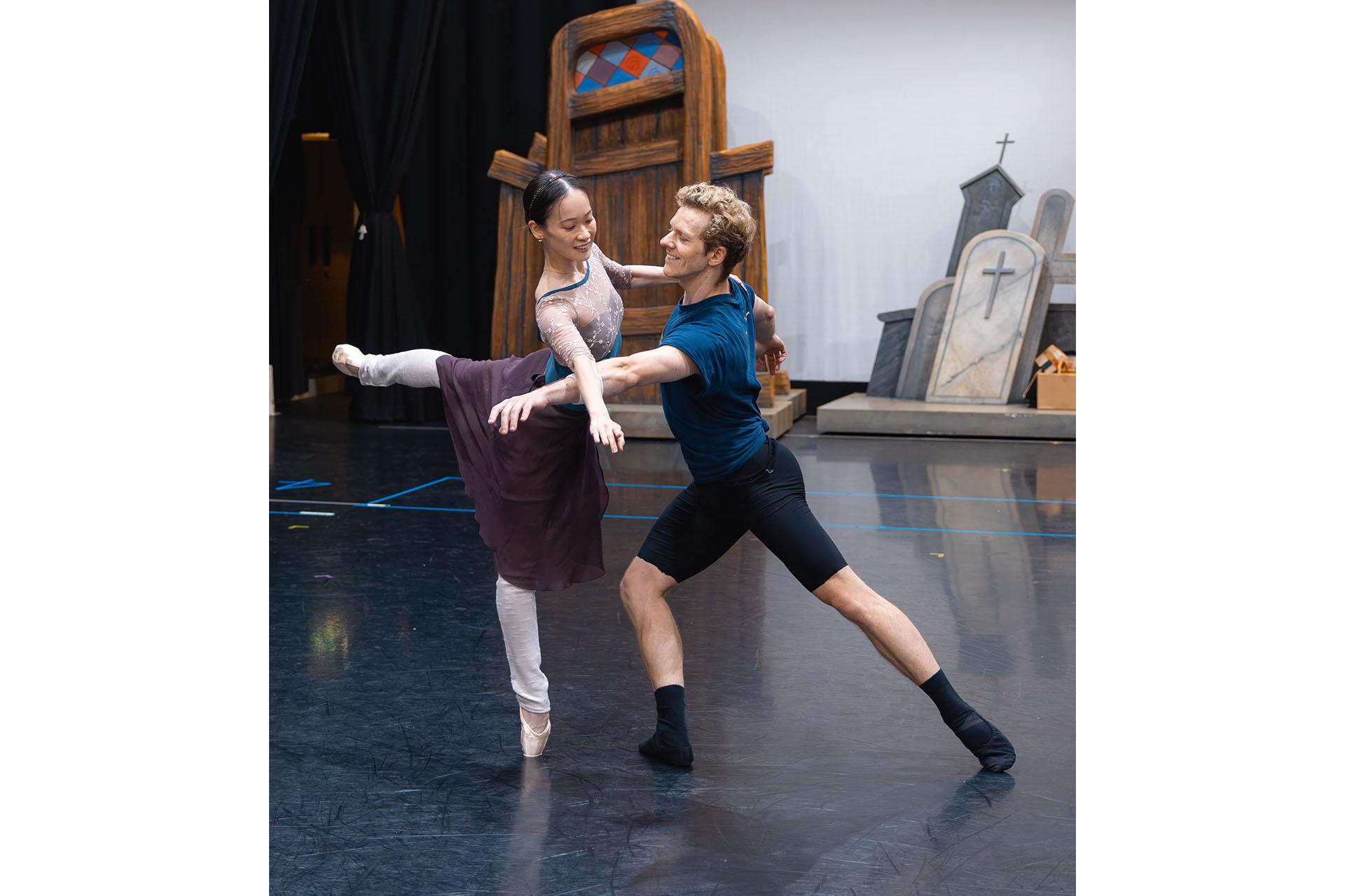 In rehearsal a dancer en point on one leg, the other raised behind her, the male dancer stands in front of her, his arms beneath hers helping her balance