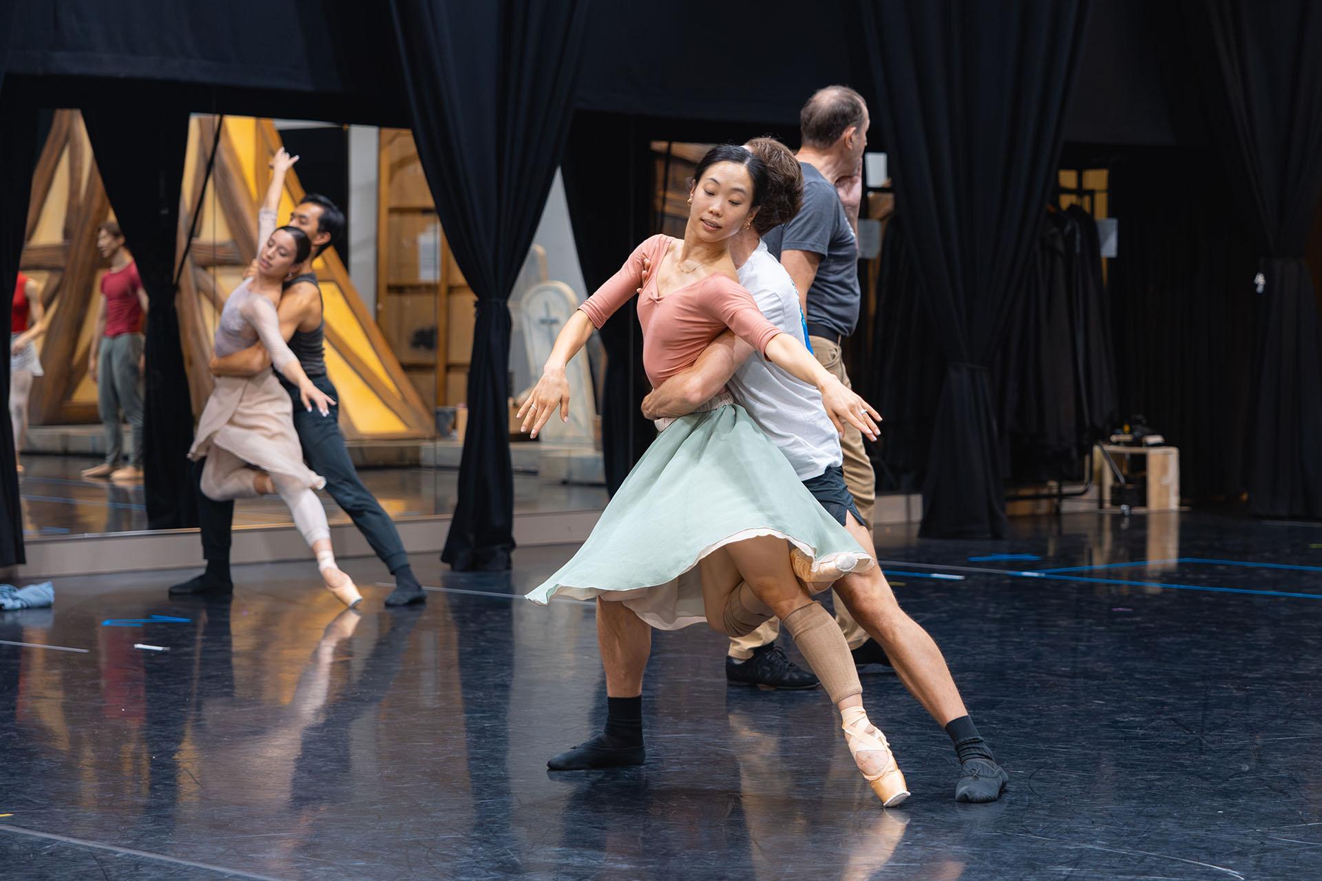 A dancer leans forward, one leg behin the knee of the other, her skirt flared with movement, an arm around her waist carrying her forward