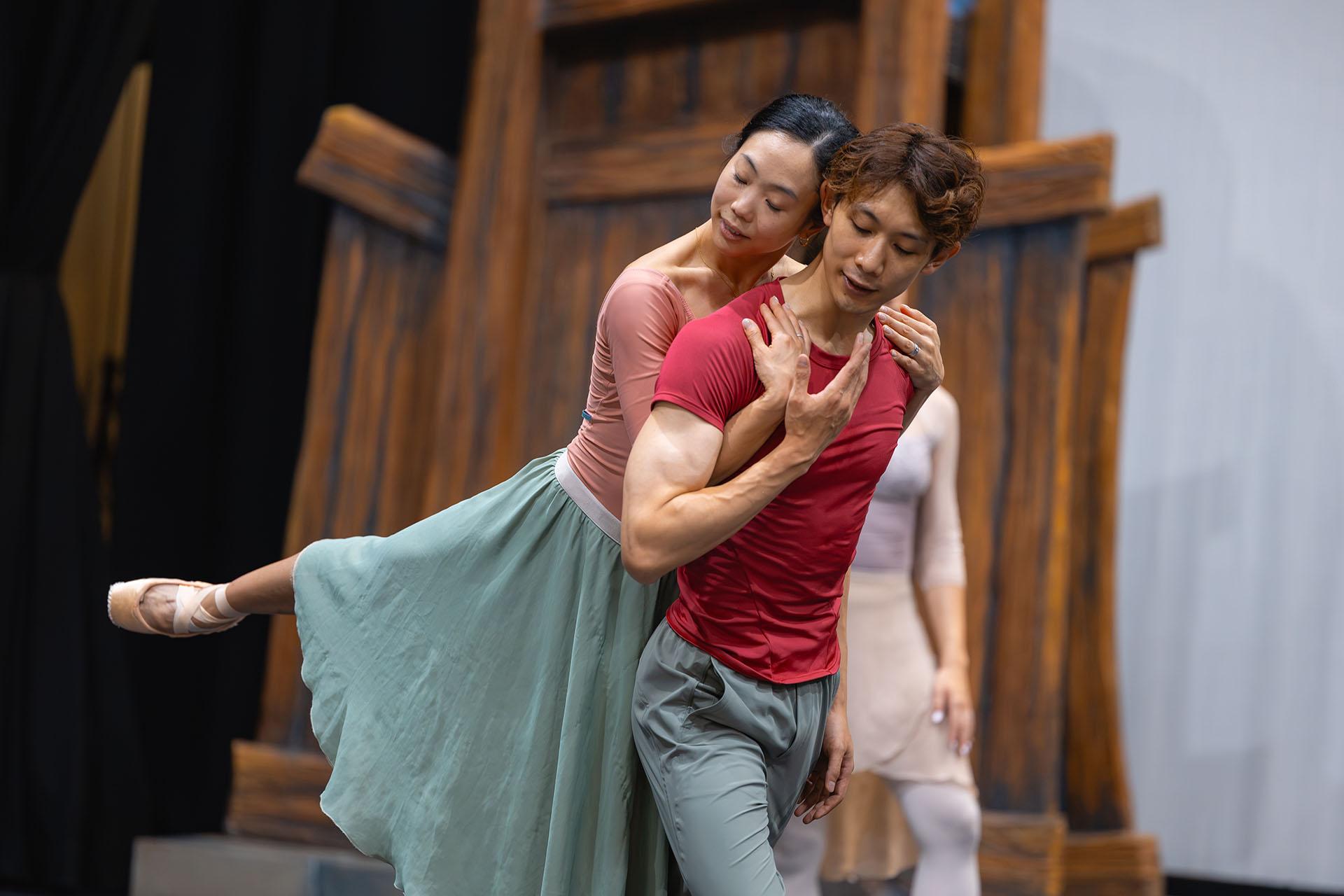 Dancer in a red t-shirt move forward, another holds him from behind and is lifted as she hooks her arms under his and holds his shoulders