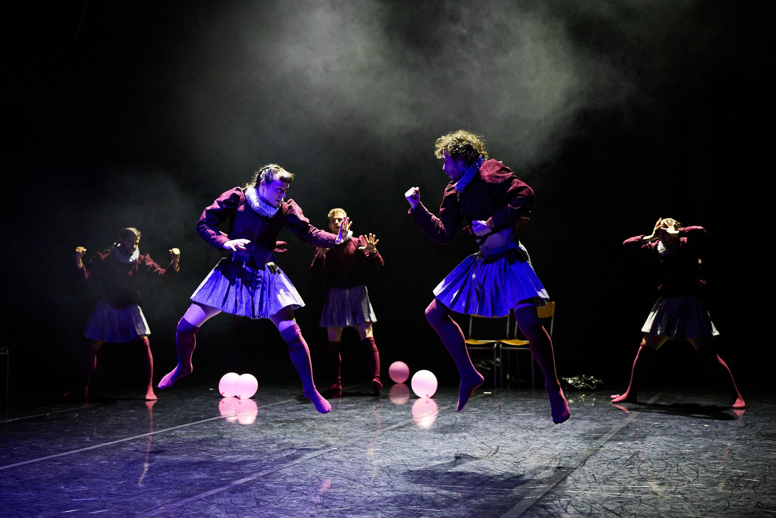 Two dancers are posed in a 'fight' position with their hands in fists as they both jump into the air. There is a crowd of other dancers behind them.