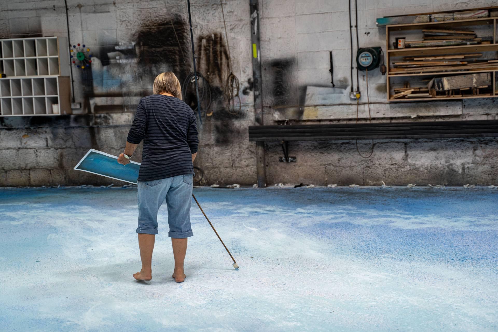 A person wearing a black top and jeans that are rolled up at the hems stands on a large canvas painted in shades of blue. They are holding a large paintbrush on one hand and a reference image on the other as they paint the canvas.