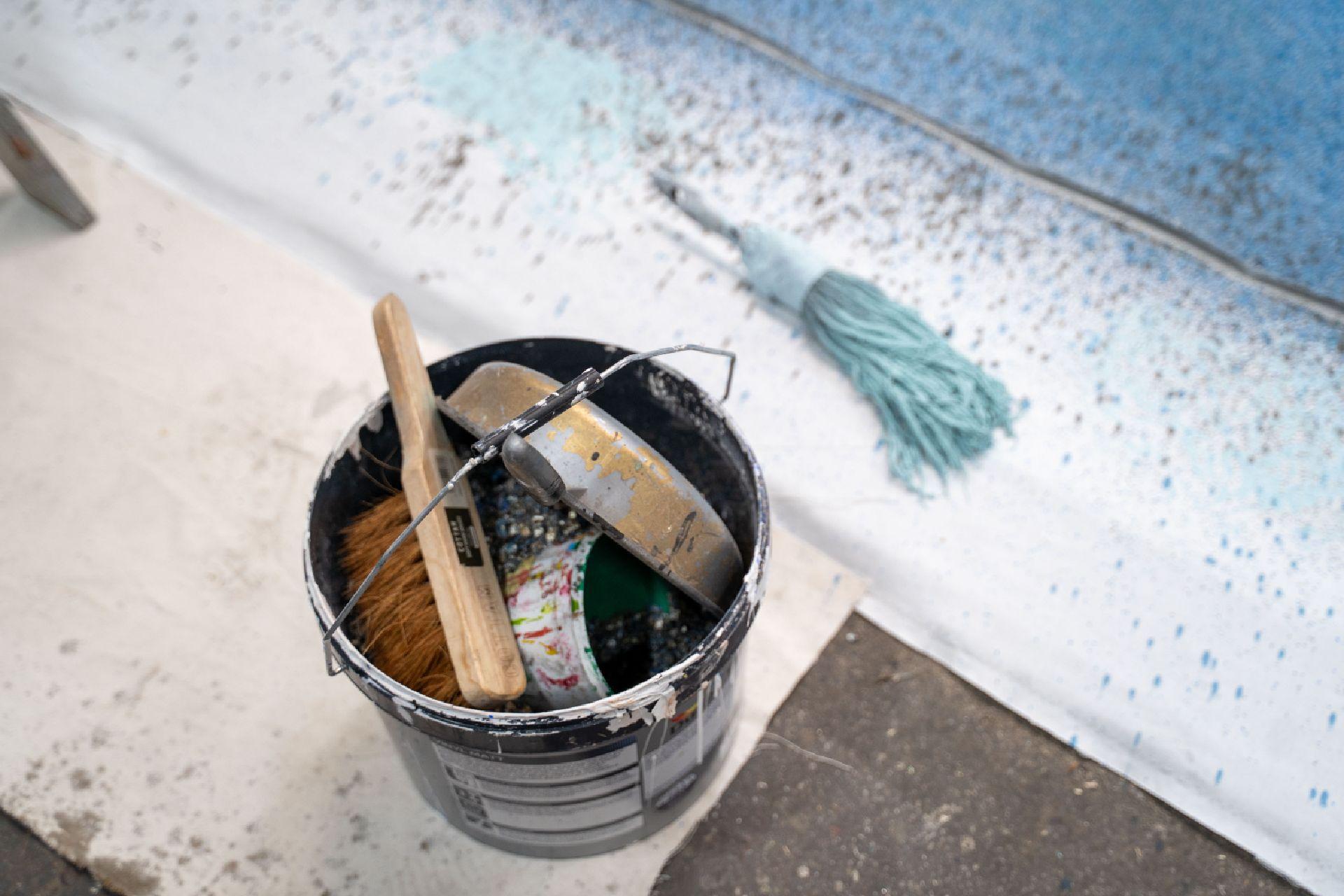 A black bucket with painting tools and utensils placed on the floor. Next to it lies a large paintbrush covered in blue paint.