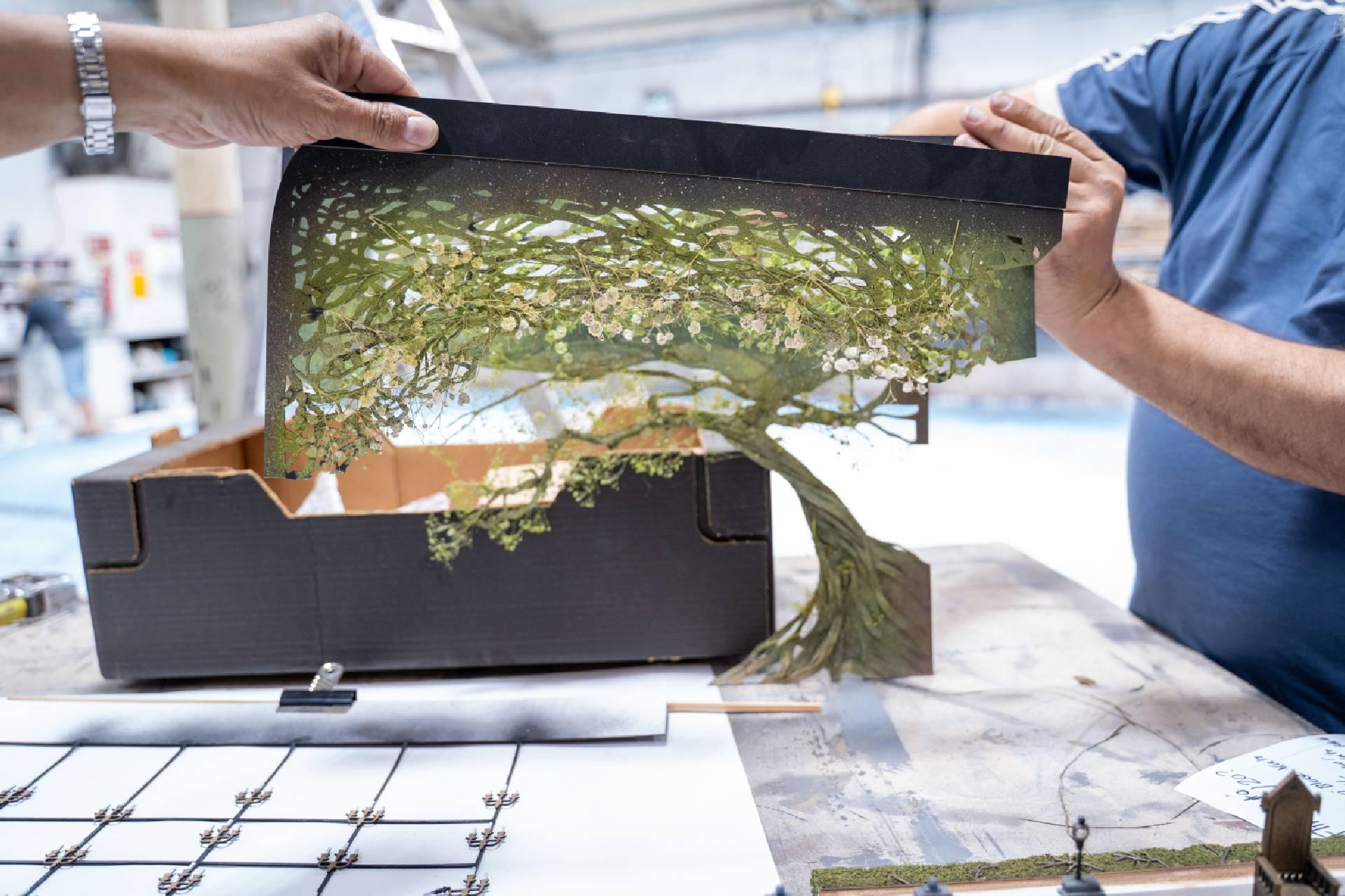 A person in a blue shirt holds a model of a windswept tree covered in green moss.