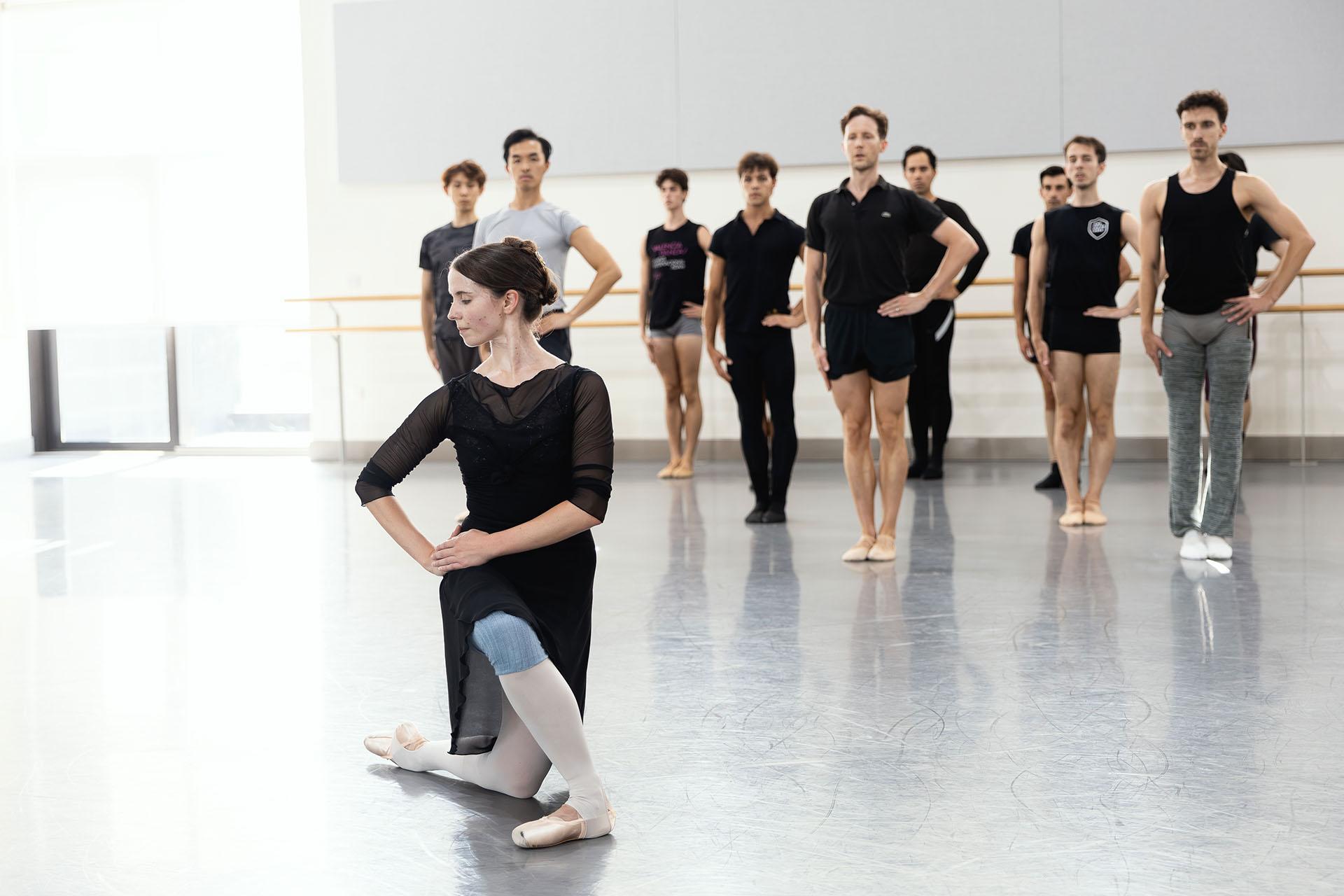 Dancers kneels on the floor, both hands on a hip, she looks sideways and down, behind her a groups of male dancers - each with a hand on their left hip, their right hand straight down