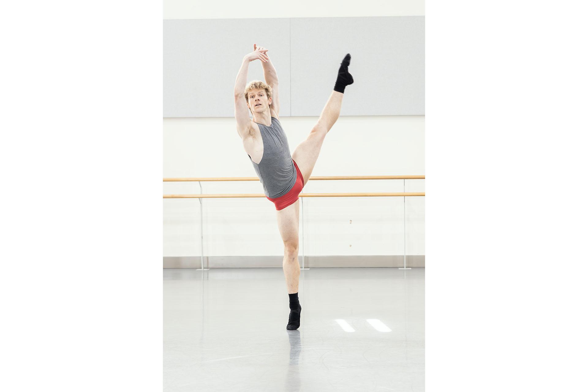 Dancer at the height of a dévelloppé à la seconde, wearing red shorts, black ballet shoes and grey vest.