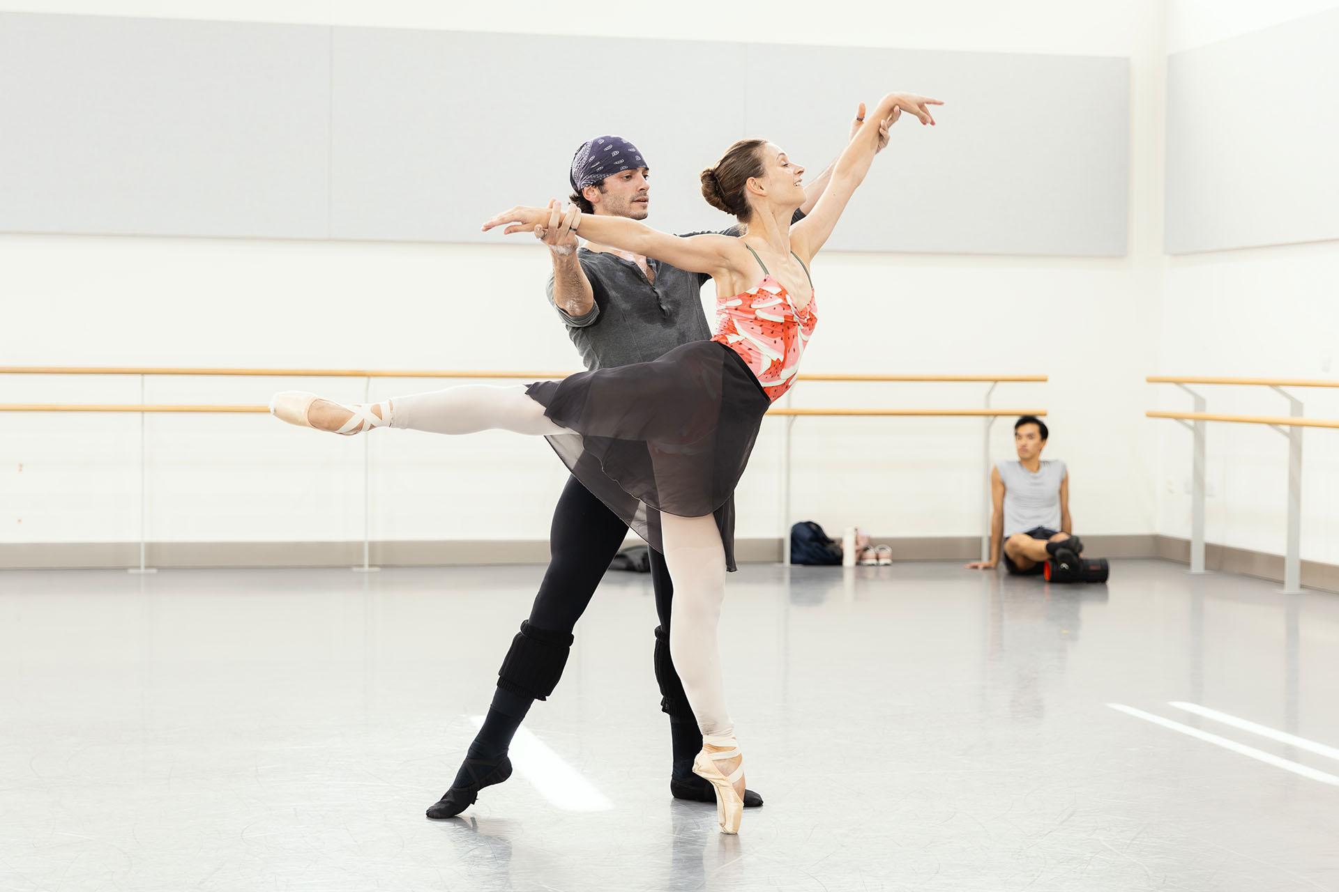 A dancer en pointe in arabesque, another dancer behind her holding her arms steady by supporting her below her wrists