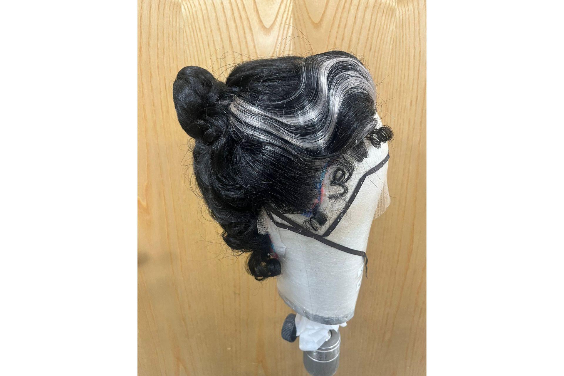 A black wig with a grey stripe has been curled. It is pictured in profile.