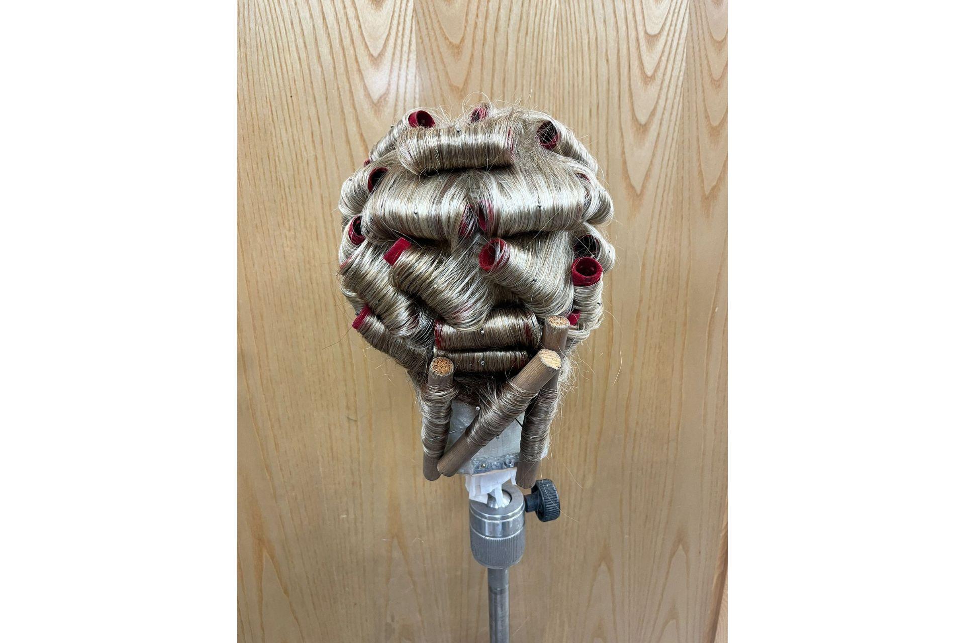 A blonde wig is fitted with red rollers in the making process. They are curled very tight and there are also wooden rollers at the bottom. It is pictured from behind.